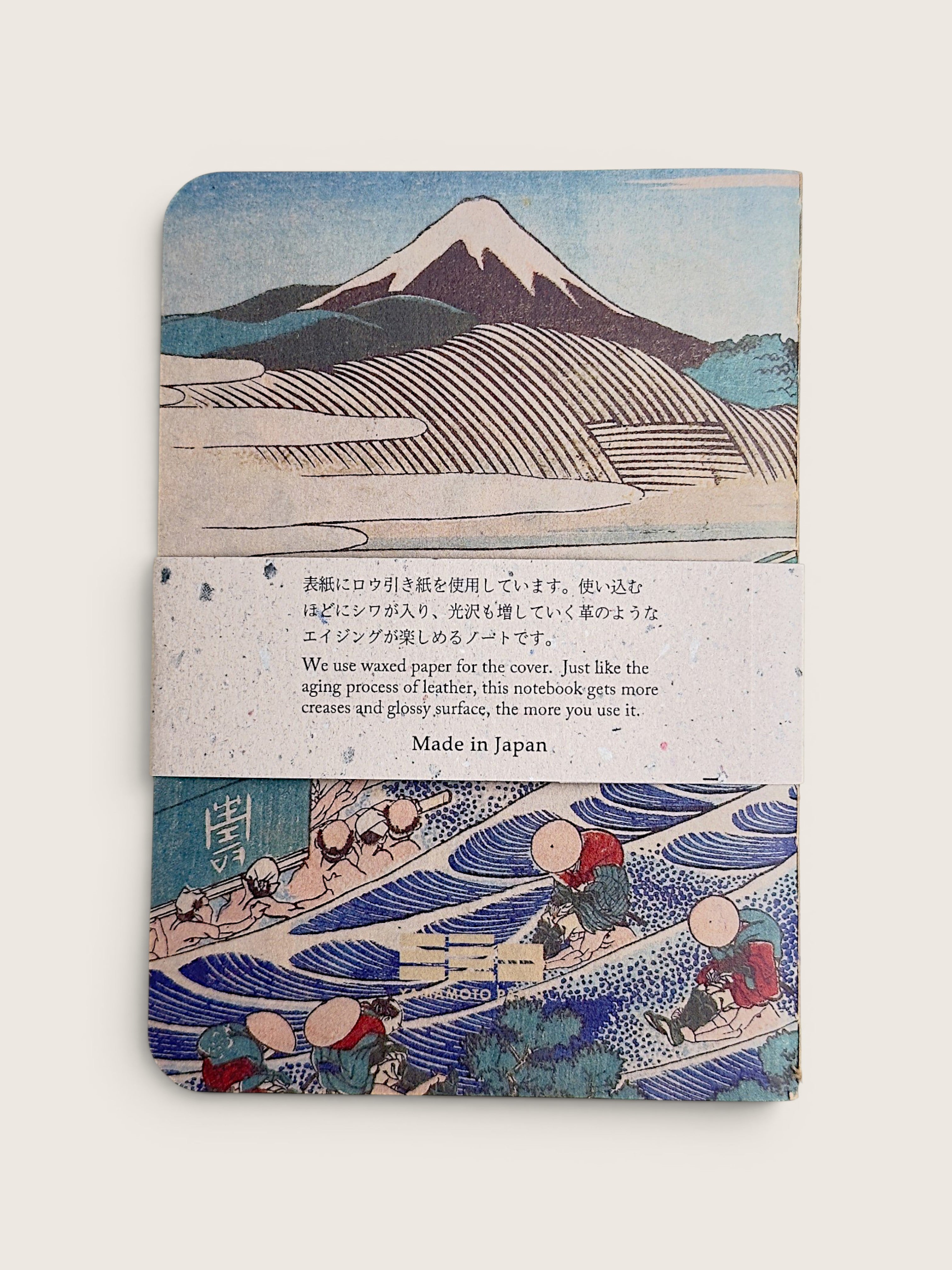 A vintage-style notebook or journal with a Japanese woodblock print cover featuring Mount Fuji and waves, wrapped with a paper band that reads 'Made in Japan' with additional text about wound paper.