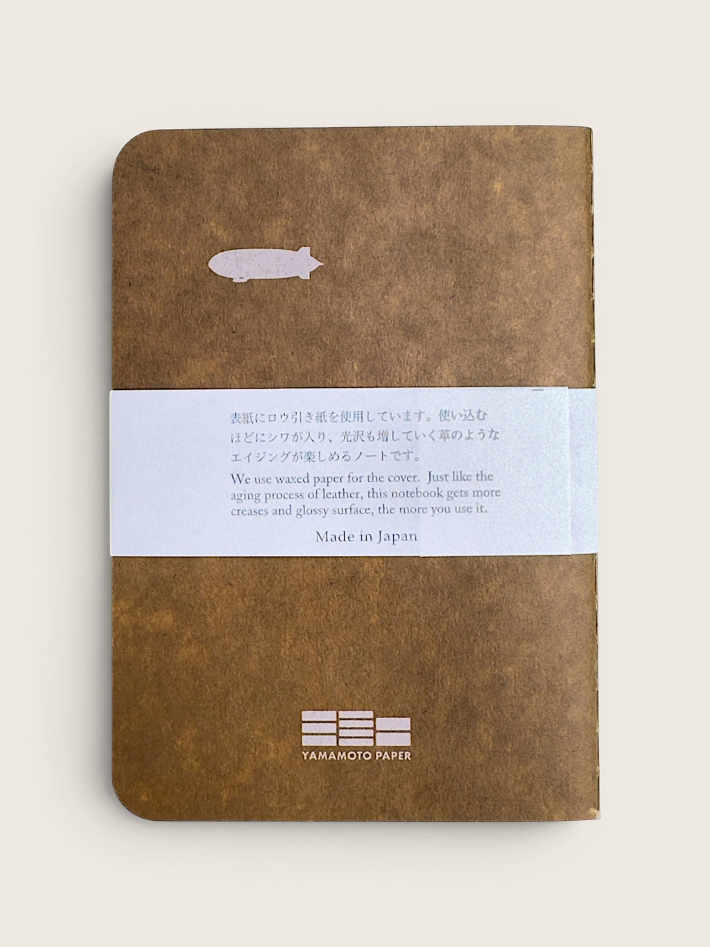 A brown textured notebook with a white paper band wrapped around the middle, photographed on a light beige background. The notebook has a small white fish illustration near the top and the text 'YAMANIZTO PAPER' printed at the bottom.