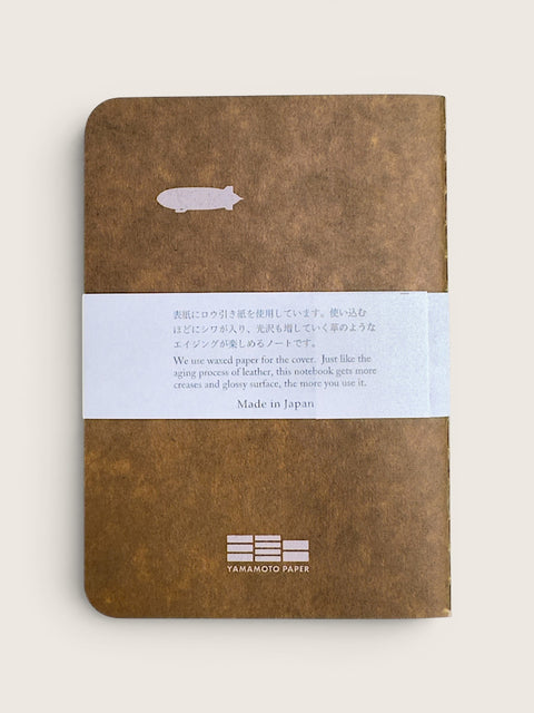 A brown textured notebook with a white paper band wrapped around the middle, photographed on a light beige background. The notebook has a small white fish illustration near the top and the text 'YAMANIZTO PAPER' printed at the bottom.