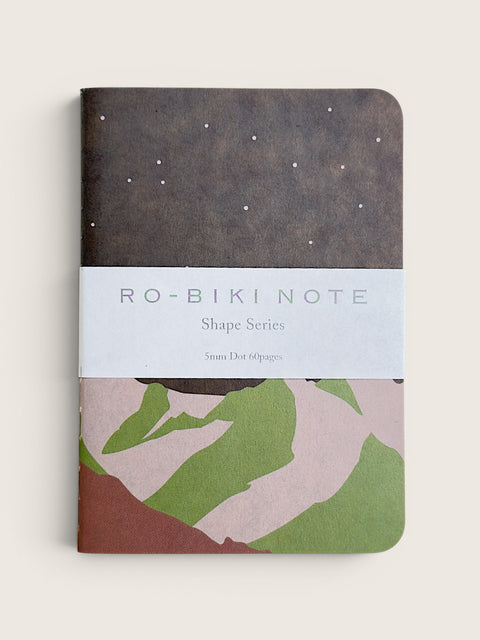 A notebook from RO-BIKI NOTE Shape Series, featuring a dark brown textured top half with white star-like dots, a white paper band across the middle with the brand name, and a bottom half with abstract green, pink, and brown shapes, shown on a light beige background.