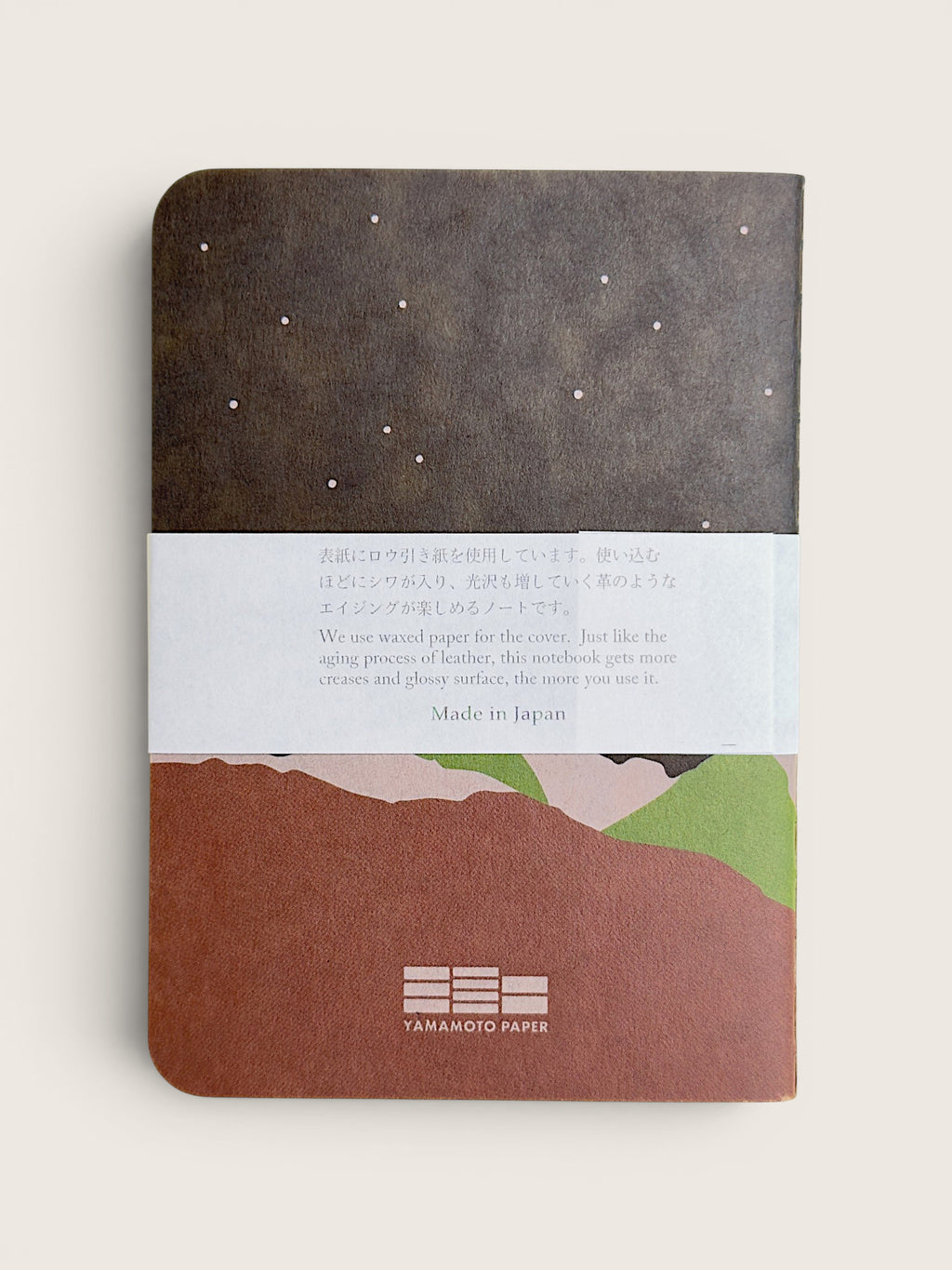 A Yamamoto Paper notebook with a dark brown top section featuring white stars, a white paper band with Japanese and English text, and a bottom section showing a brown, pink, and green landscape illustration.