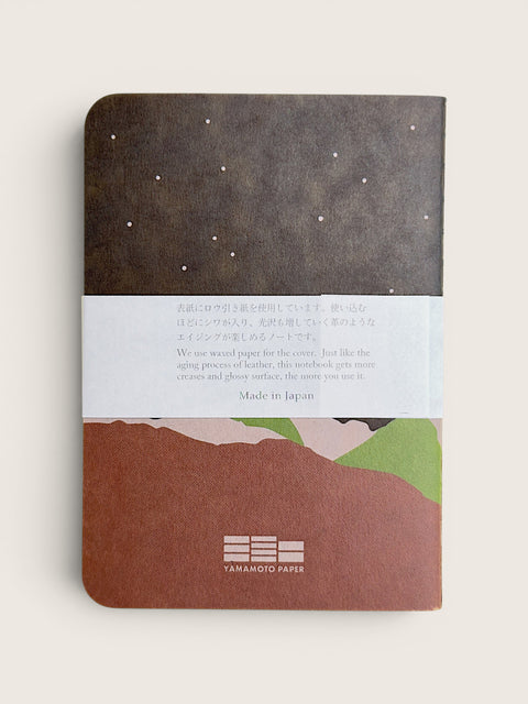 A Yamamoto Paper notebook with a dark brown top section featuring white stars, a white paper band with Japanese and English text, and a bottom section showing a brown, pink, and green landscape illustration.