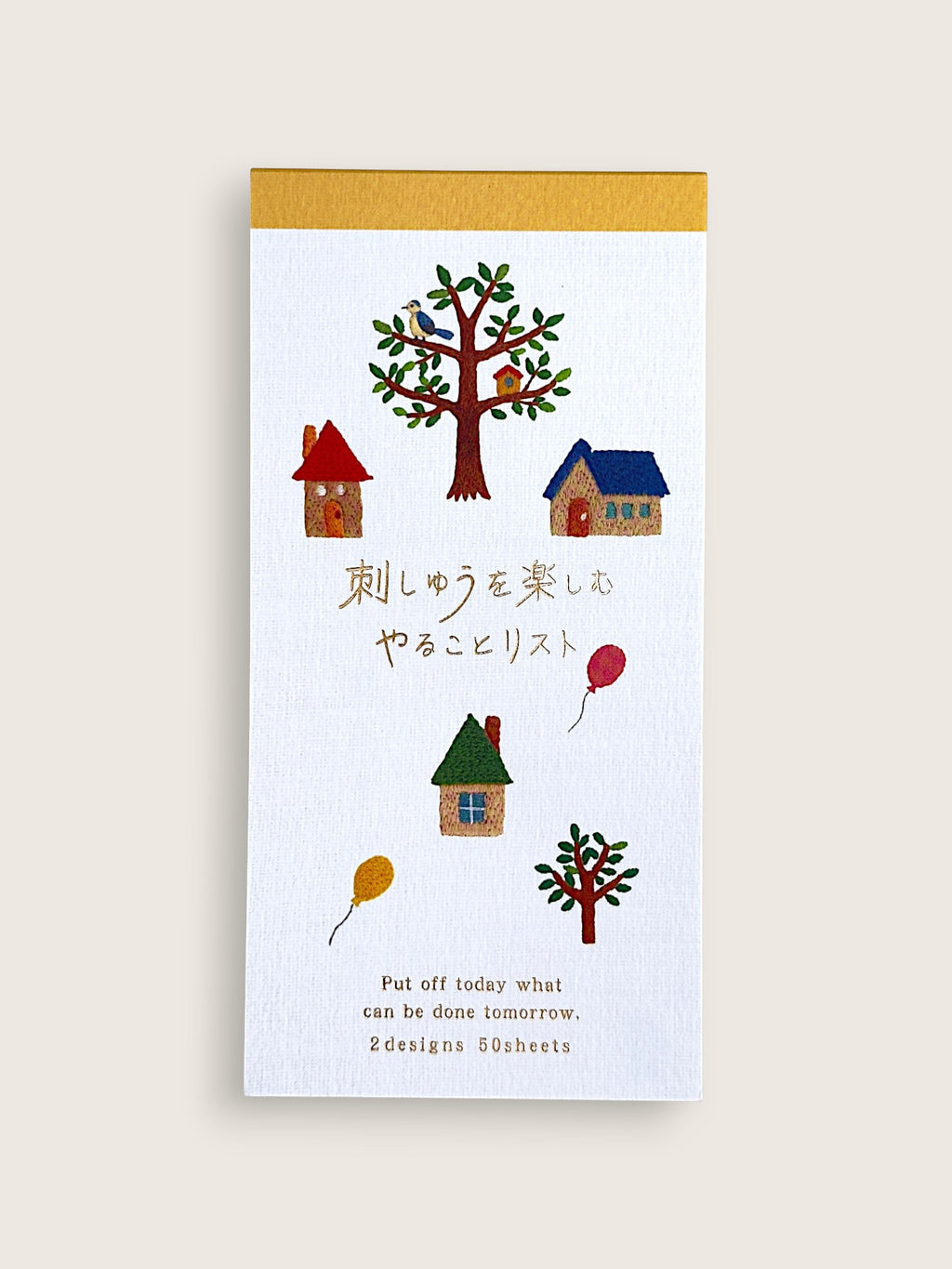 A vertical white notepad with a gold top border, decorated with small colorful illustrations of houses, trees, birds, and balloons, plus Japanese and English text. The notepad is photographed against a plain light beige background.