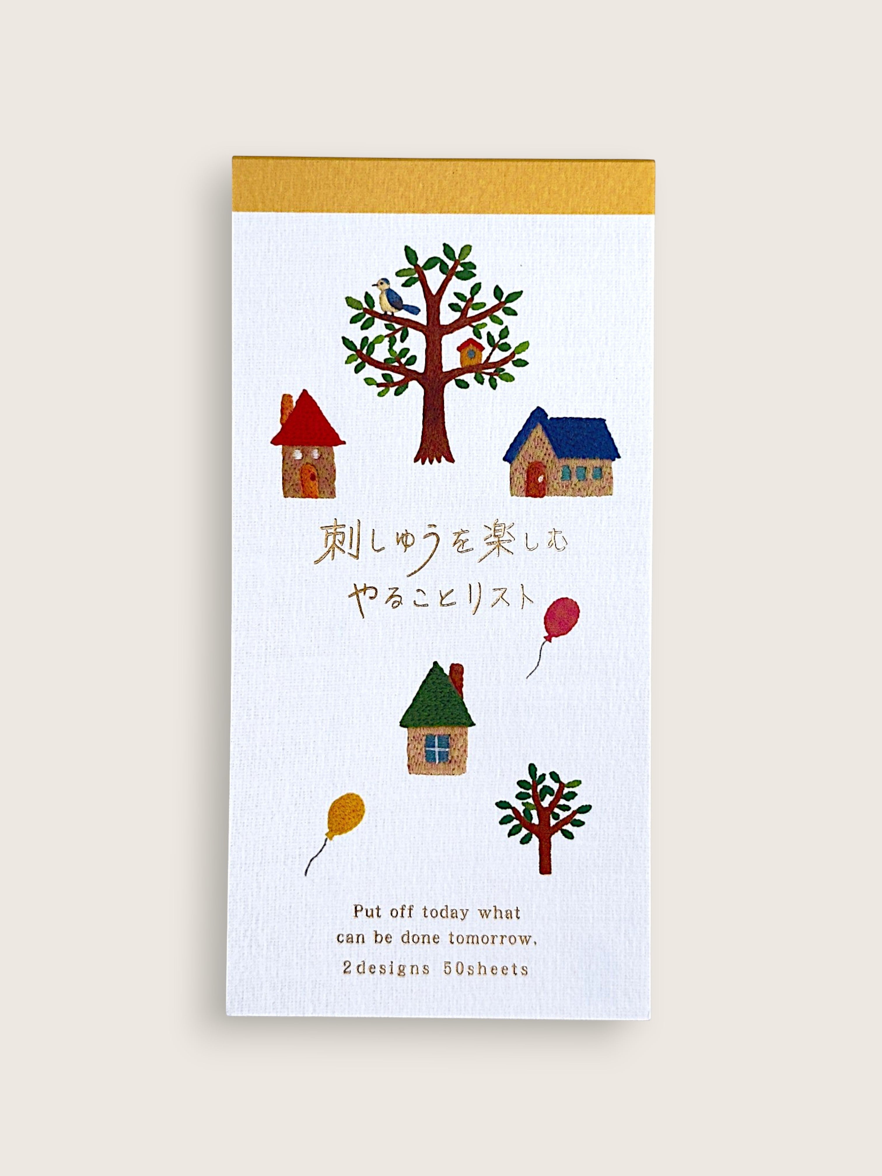 A vertical white notepad with a gold top border, decorated with small colorful illustrations of houses, trees, birds, and balloons, plus Japanese and English text. The notepad is photographed against a plain light beige background.