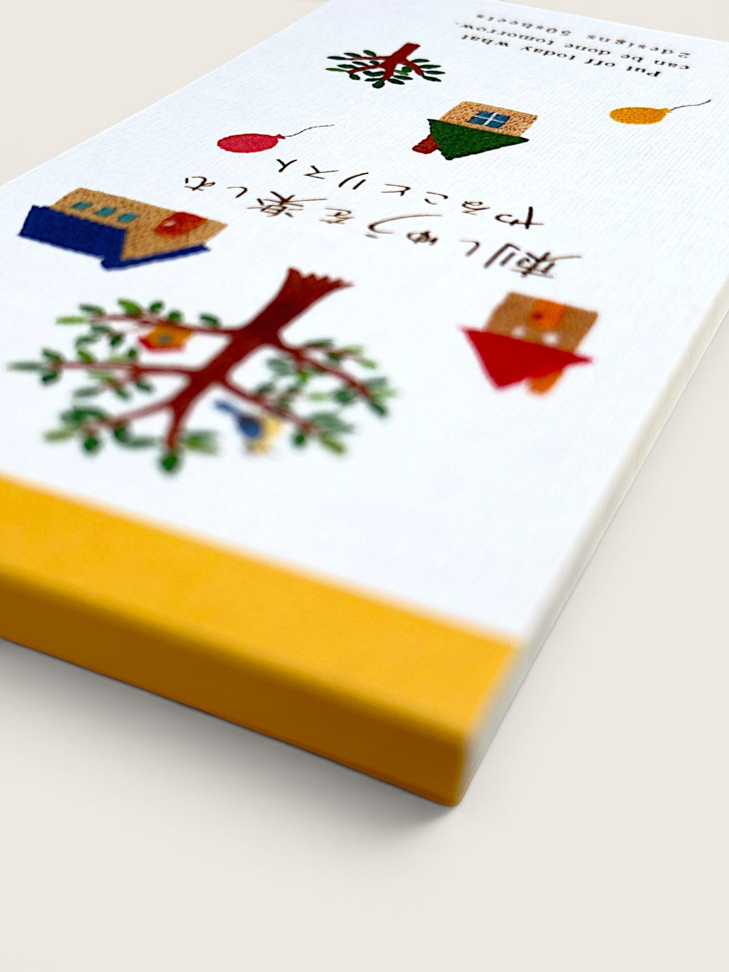 A close-up of a book or notebook cover with a white top surface decorated with small colorful illustrations and Japanese text, plus a wide yellow band along the bottom edge, photographed on a light neutral background.