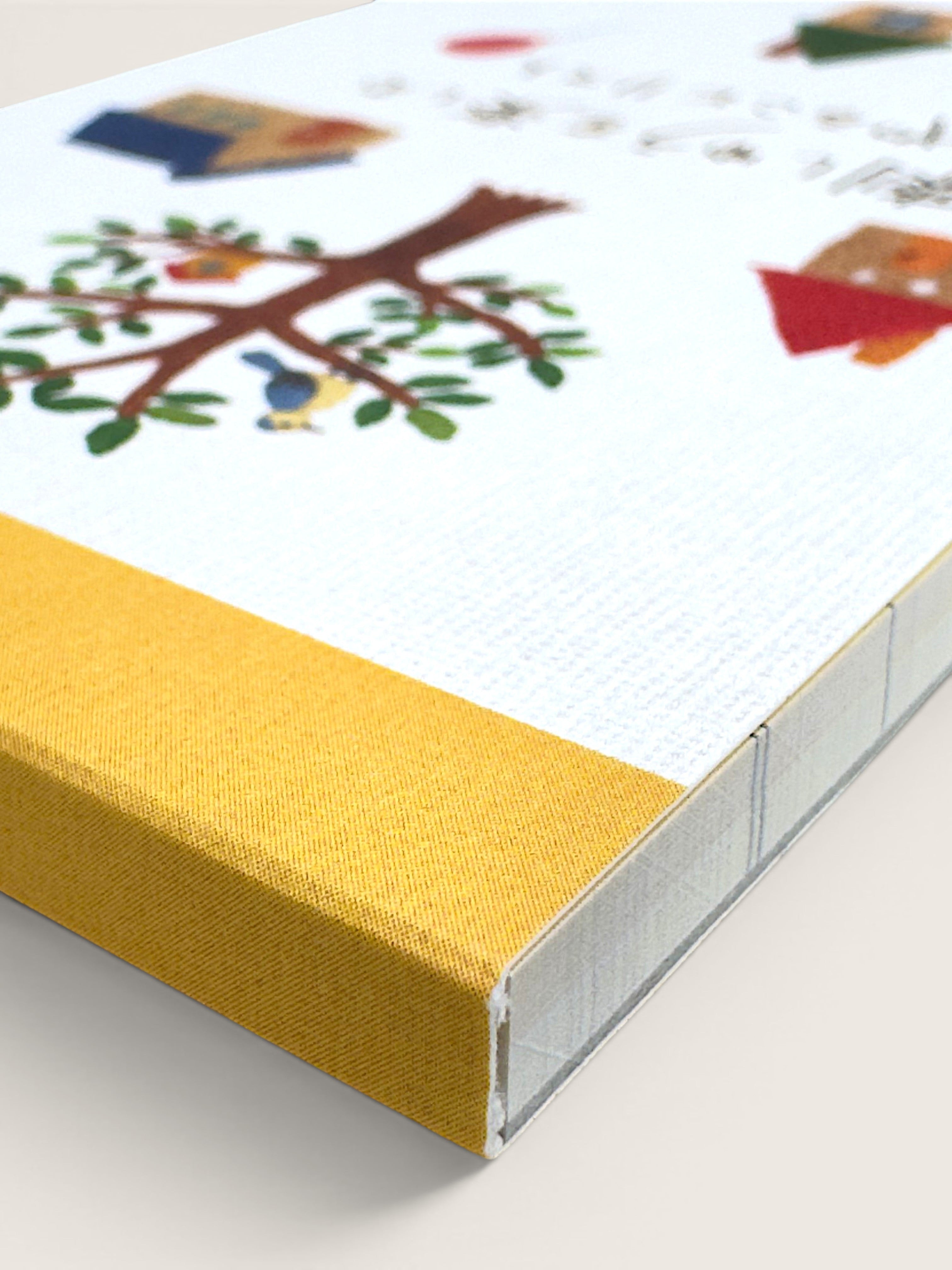 A close-up of a hardcover book or notebook with a white cover decorated with colorful illustrations of a tree, birds, and small houses, bound with a mustard yellow fabric spine.