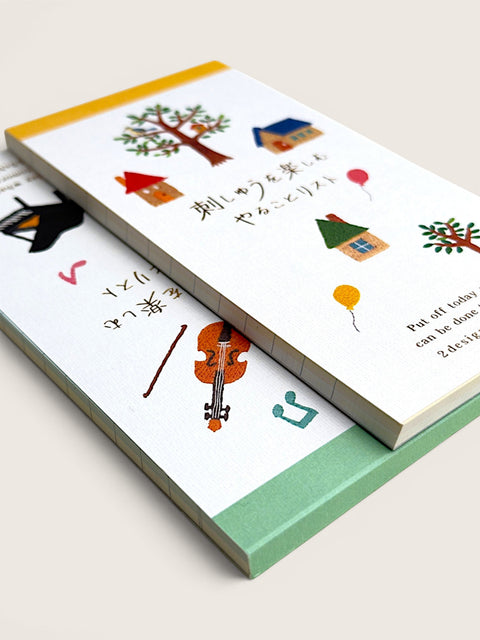 Two illustrated notebooks or books stacked at an angle on a light beige background. The top notebook has a white cover with colorful drawings of trees, houses, balloons, and Japanese text, plus a partial English quote.