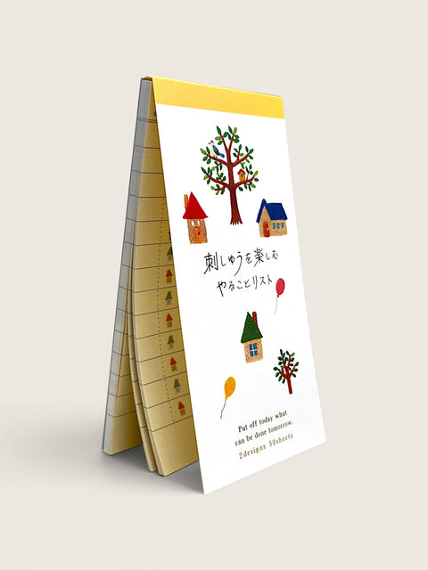 A freestanding A-frame paper planner or checklist with a white front cover decorated with colorful illustrations of trees, houses, and balloons, plus Japanese text and a small English phrase near the bottom. The interior pages are beige with printed measurement or task lines.