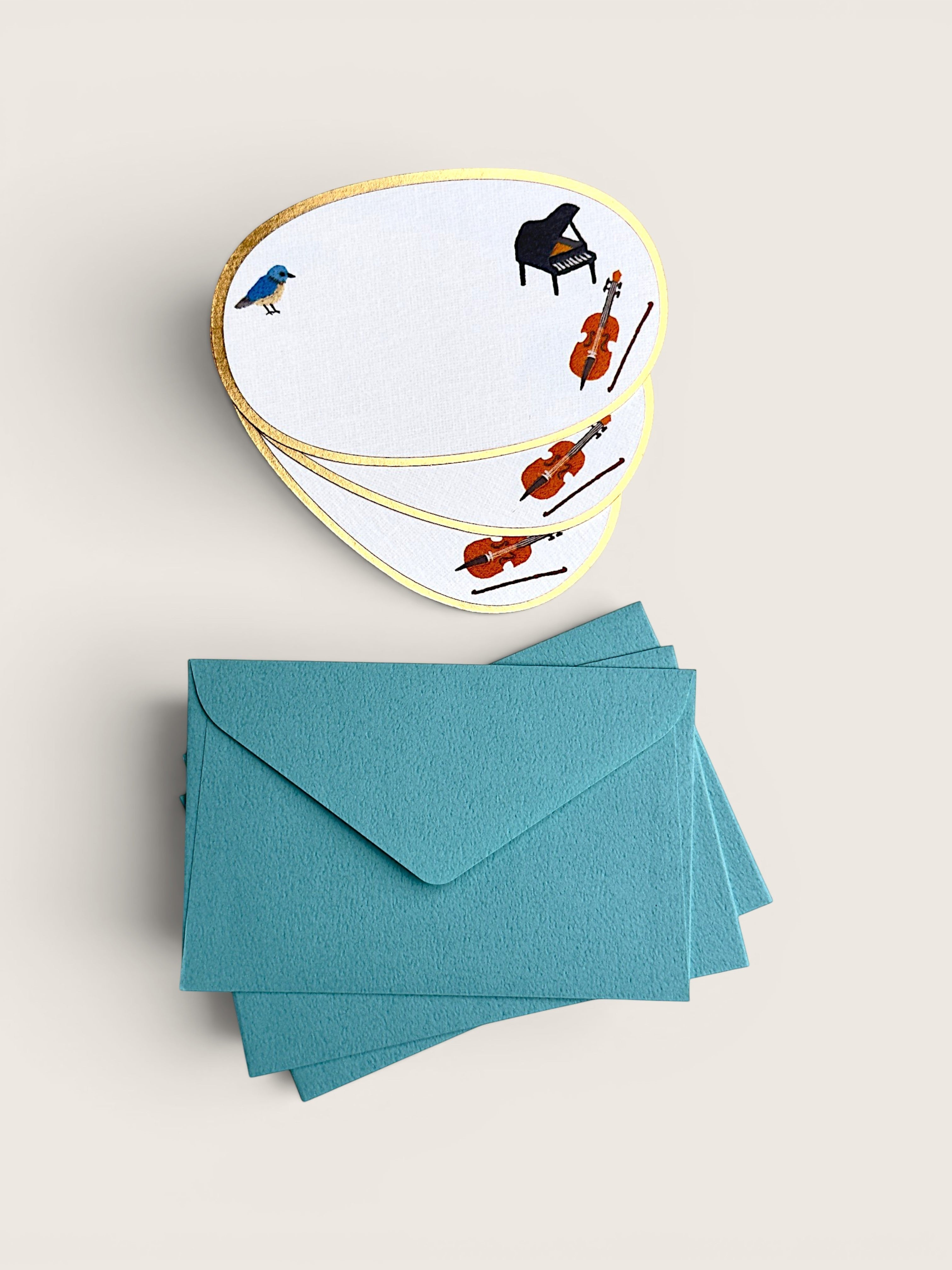 A stack of white guitar-shaped cards with gold edges, decorated with small illustrations of a bird, piano, cello, and violins, shown above a matching stack of teal envelopes on a light beige background.