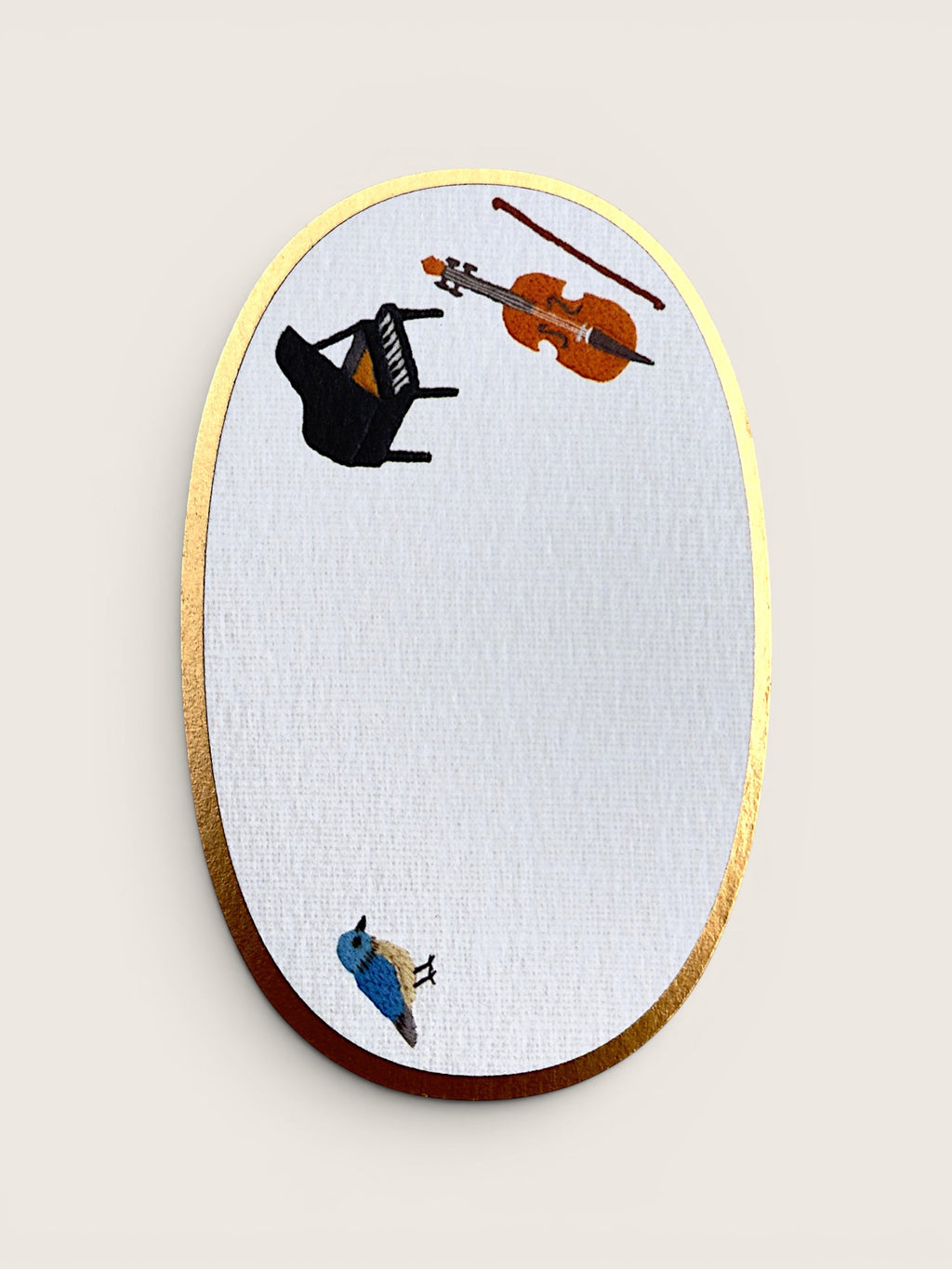 An oval wall mirror with a gold-toned metallic frame and a white textured glass surface, decorated with whimsical illustrations of a black grand piano, an orange violin, a brown bow, and a small blue bird, shown against a light beige background.