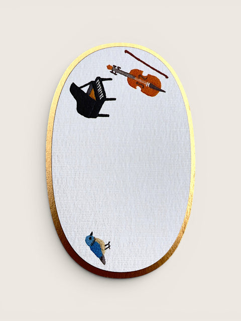 An oval wall mirror with a gold-toned metallic frame and a white textured glass surface, decorated with whimsical illustrations of a black grand piano, an orange violin, a brown bow, and a small blue bird, shown against a light beige background.