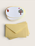 A stack of oval white stationery cards with gold borders and small whimsical illustrations, shown above a matching stack of pale yellow envelopes on a light beige background.