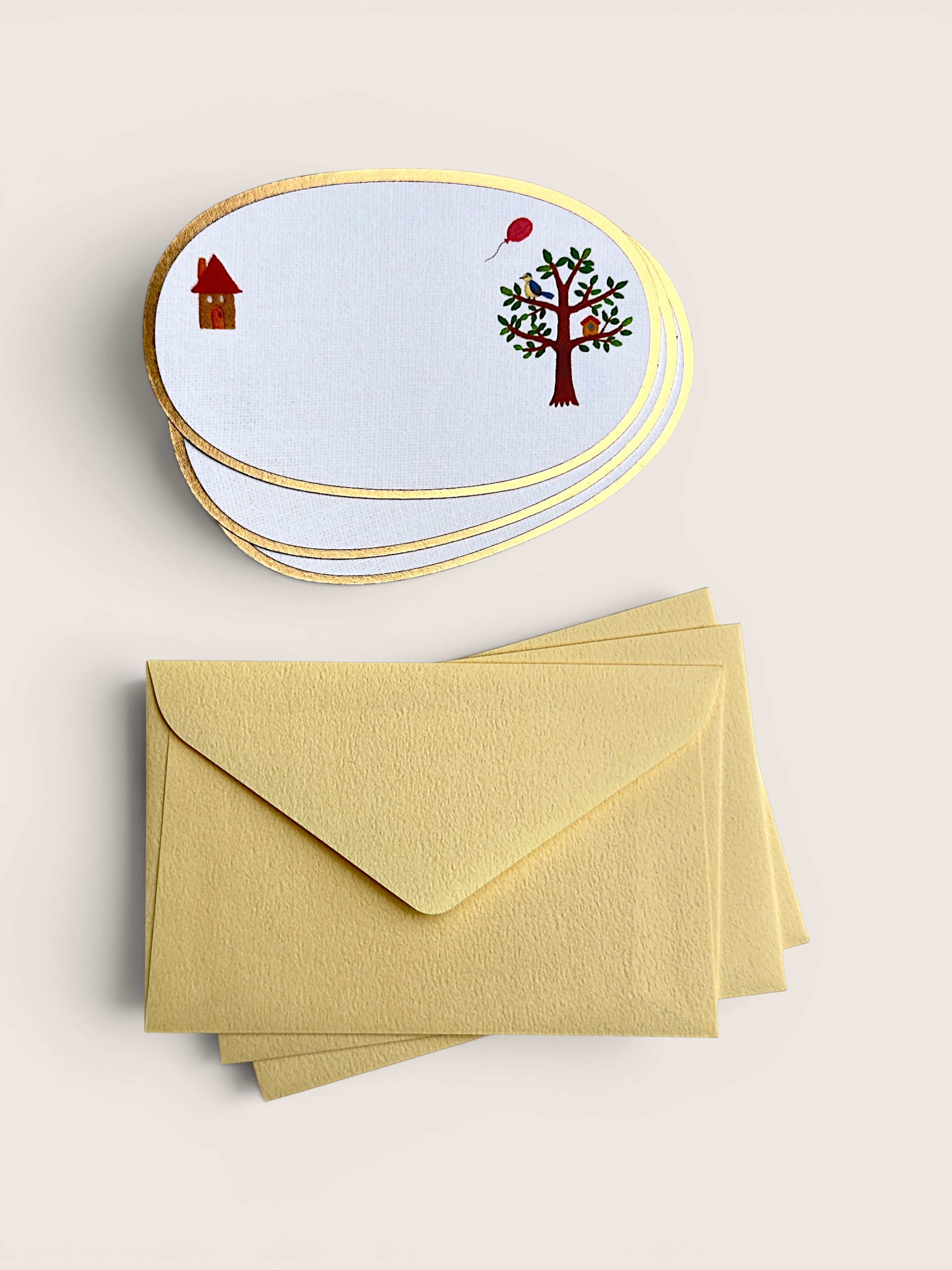 A stack of oval white stationery cards with gold borders and small whimsical illustrations, shown above a matching stack of pale yellow envelopes on a light beige background.