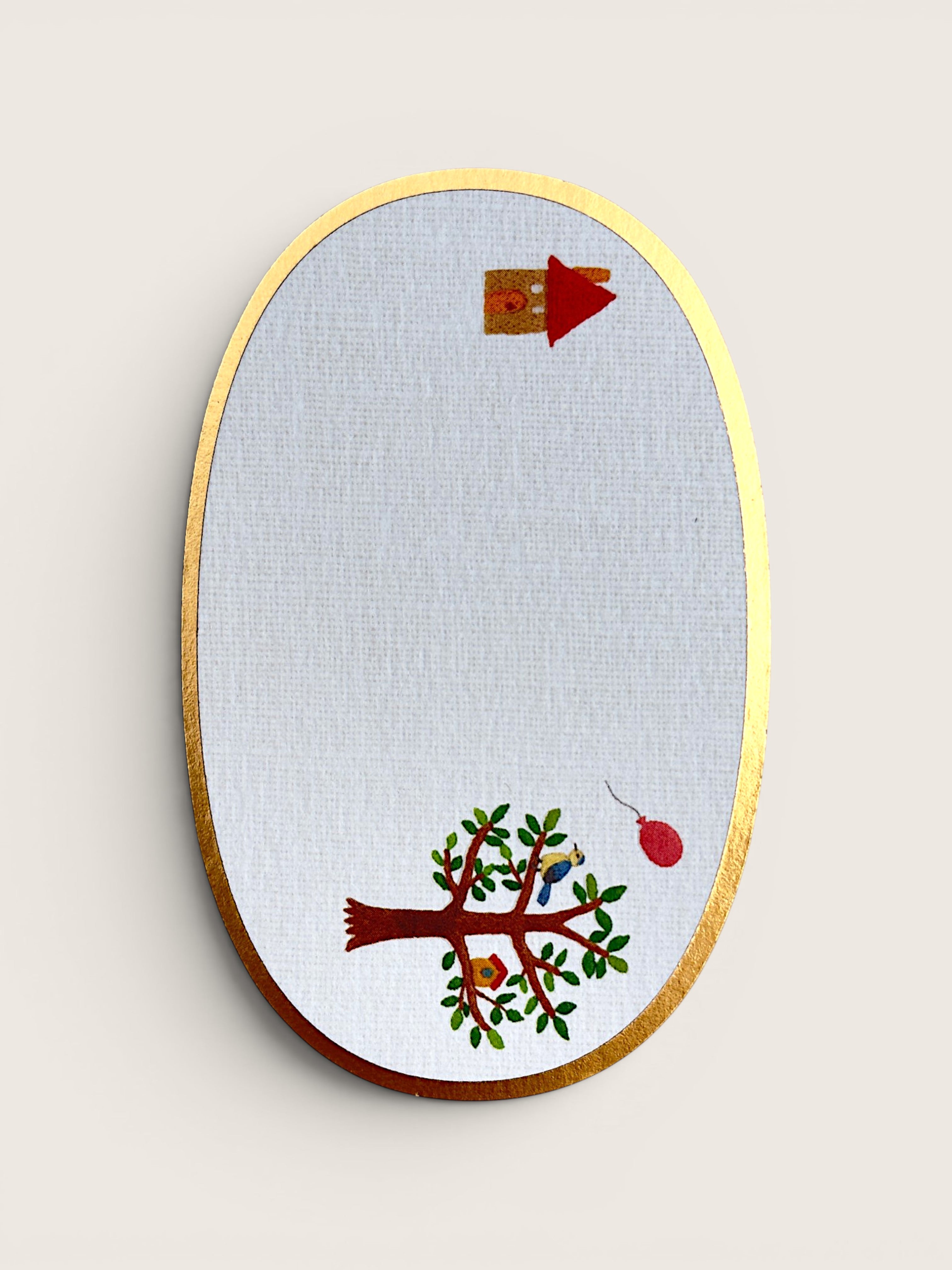 An oval wall-mounted embroidery hoop featuring a white fabric background with a gold-toned wooden frame. The embroidery design includes a brown tree with green leaves, a small birdhouse, and a red hanging ornament, set against a plain light beige wall.