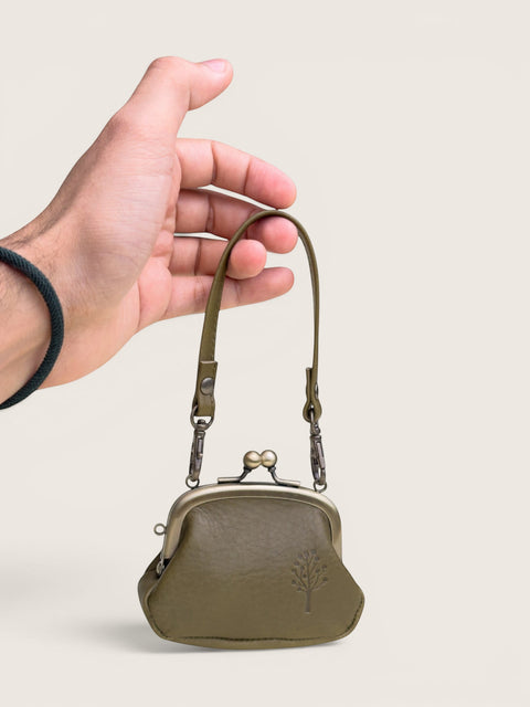 An olive green leather kiss-lock coin purse with a short shoulder strap, held by a hand against a solid light beige background.