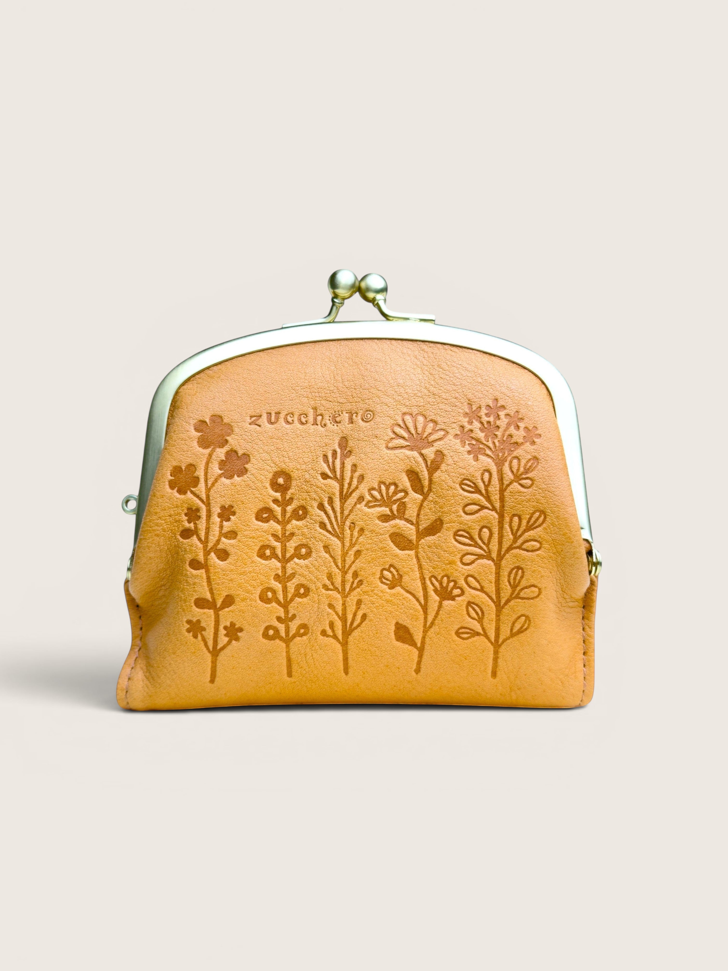 A small tan leather kiss-lock pouch with embossed floral designs and the brand name 'zucchere' printed on the front, set against a solid light beige background.