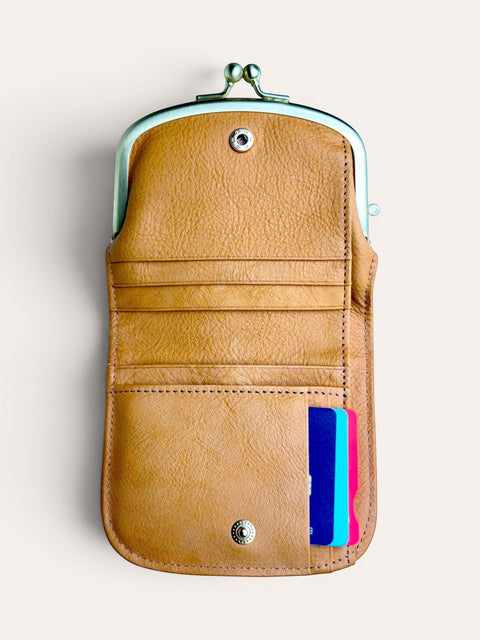 A tan leather kiss-lock wallet with a mint green metal frame and snap button, shown open with colorful credit cards in the side pocket against a light beige background.