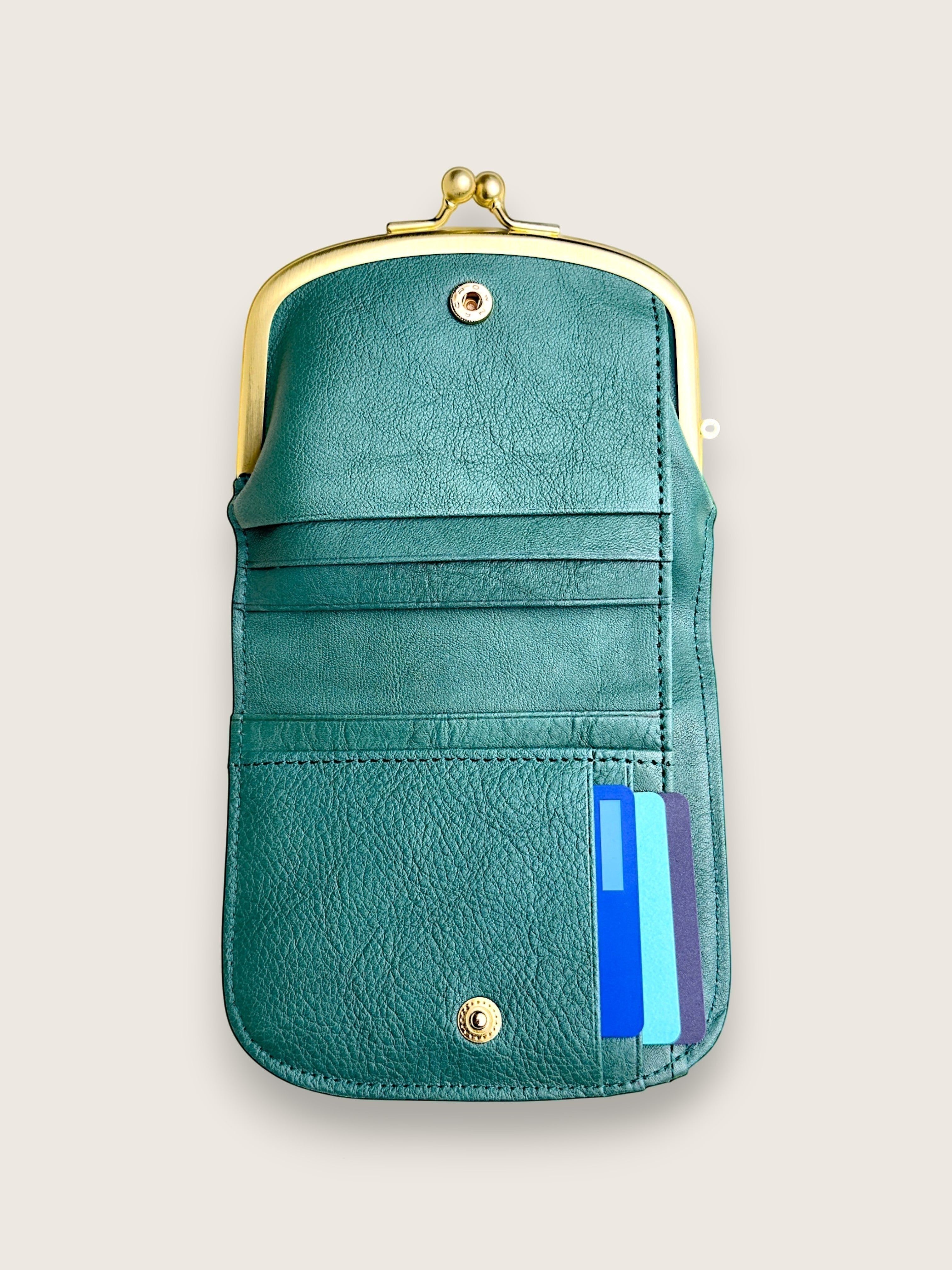 A teal green faux leather kiss-lock wallet with gold-tone metal hardware, shown open to reveal card slots and a side pocket containing blue and light blue cards, set against a solid light beige background.