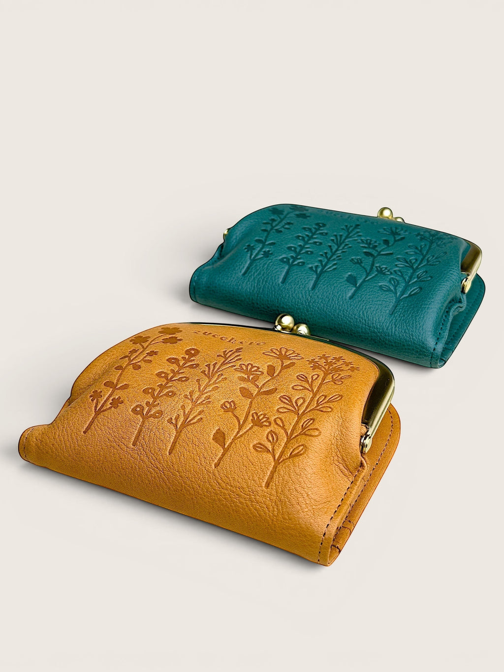 Two small leather kiss-lock wallets with embossed floral tree designs, one in mustard yellow and one in teal, shown on a light beige background.