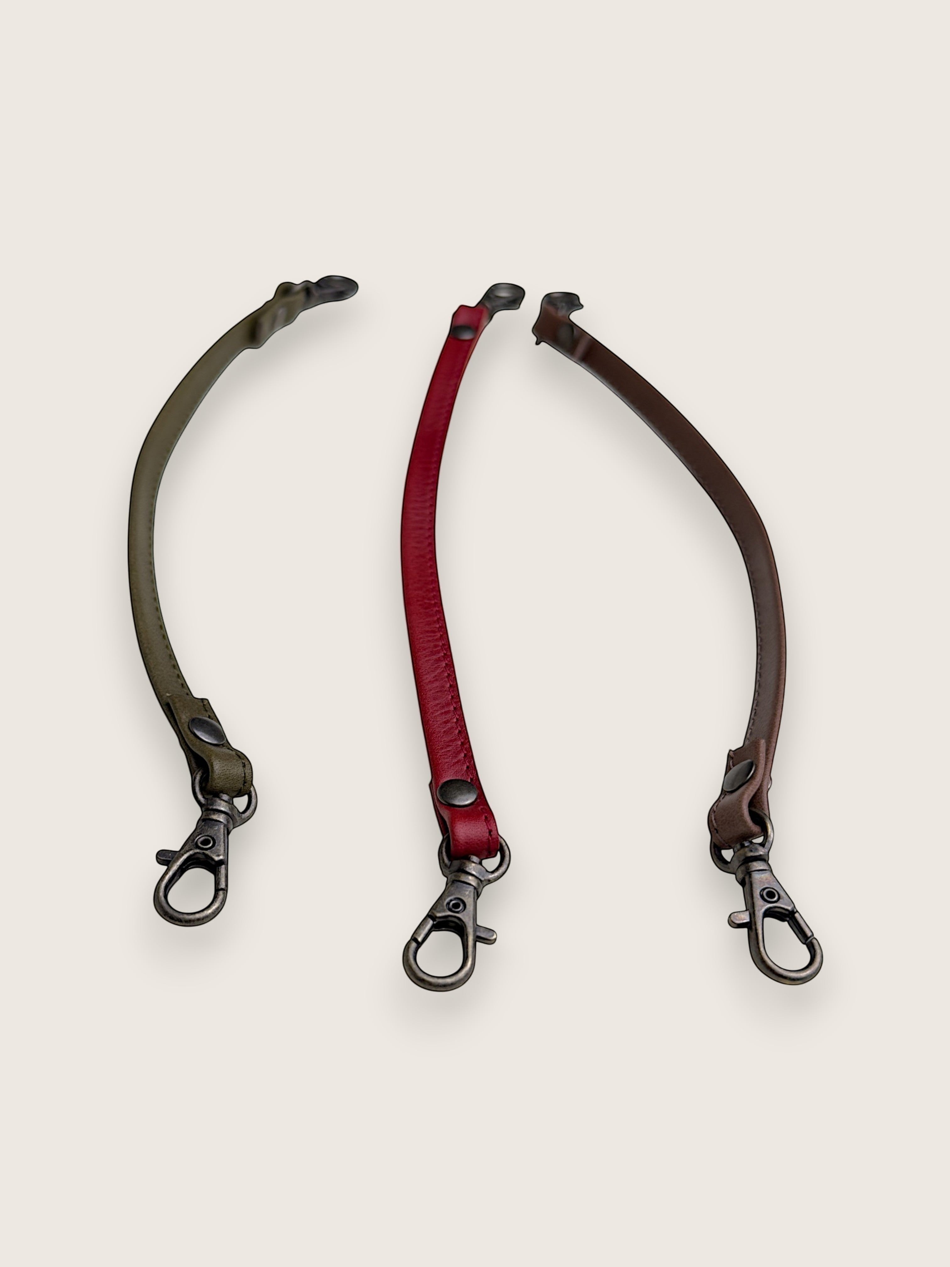 Three adjustable leather strap accessories with metal snap hooks, displayed on a light beige background. The straps are olive green, deep red, and dark brown.