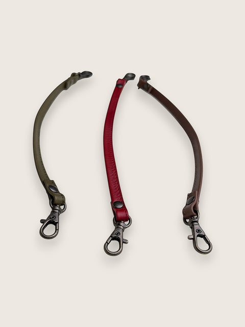 Three adjustable leather strap accessories with metal snap hooks, displayed on a light beige background. The straps are olive green, deep red, and dark brown.