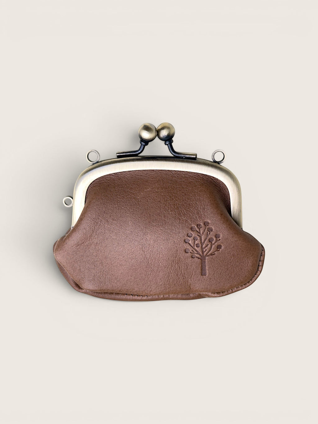 A small brown leather kiss-lock coin purse with a silver-tone metal frame and a debossed tree logo on the front, set against a solid light beige background.