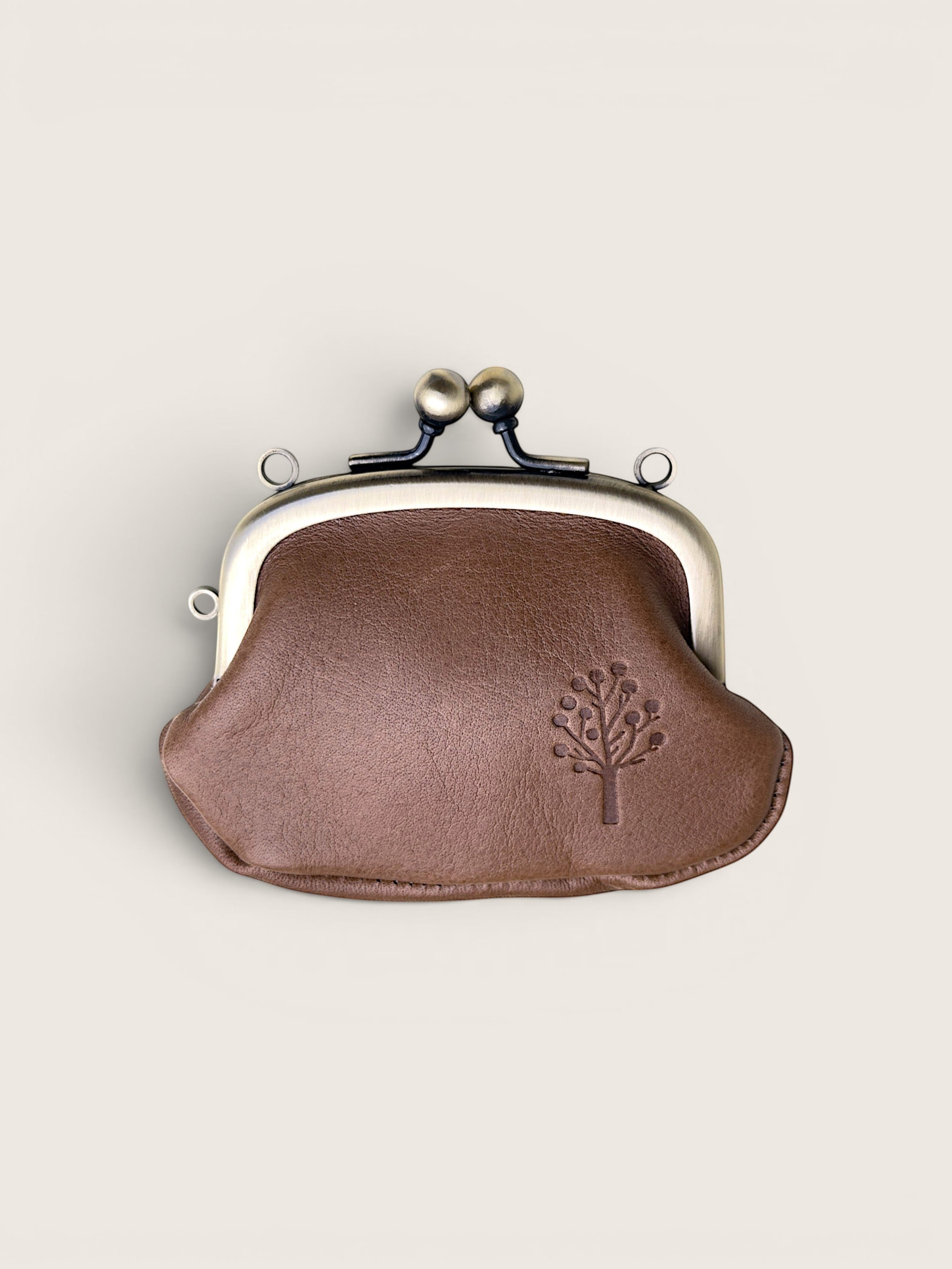 A small brown leather kiss-lock coin purse with a silver-tone metal frame and a debossed tree logo on the front, set against a solid light beige background.