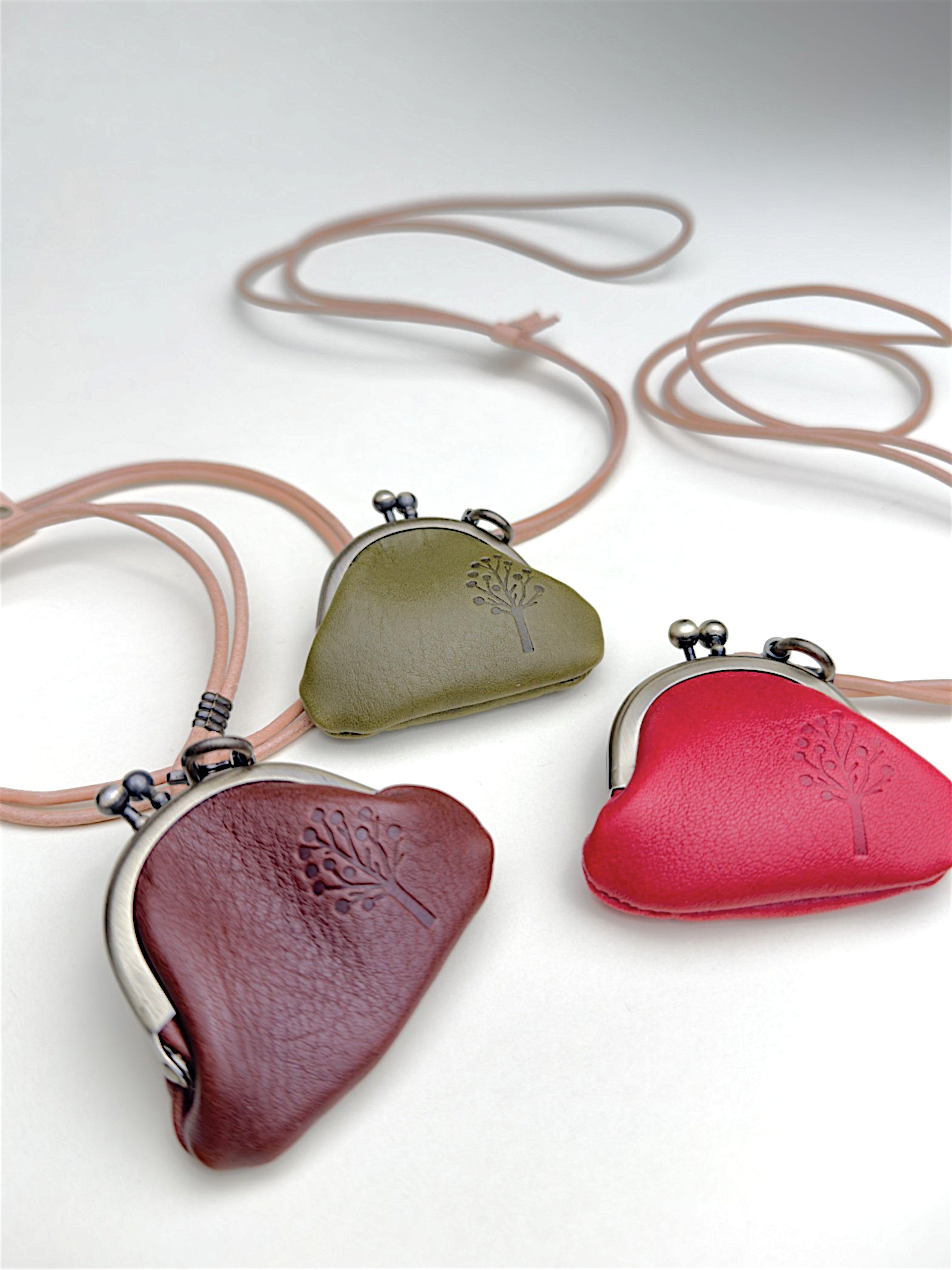 Three heart-shaped leather kiss-lock coin purses with matching necklaces, displayed on a light gray background. The purses are in burgundy, olive green, and red, with silver-tone metal frames and a small tree logo embossed on the leather.