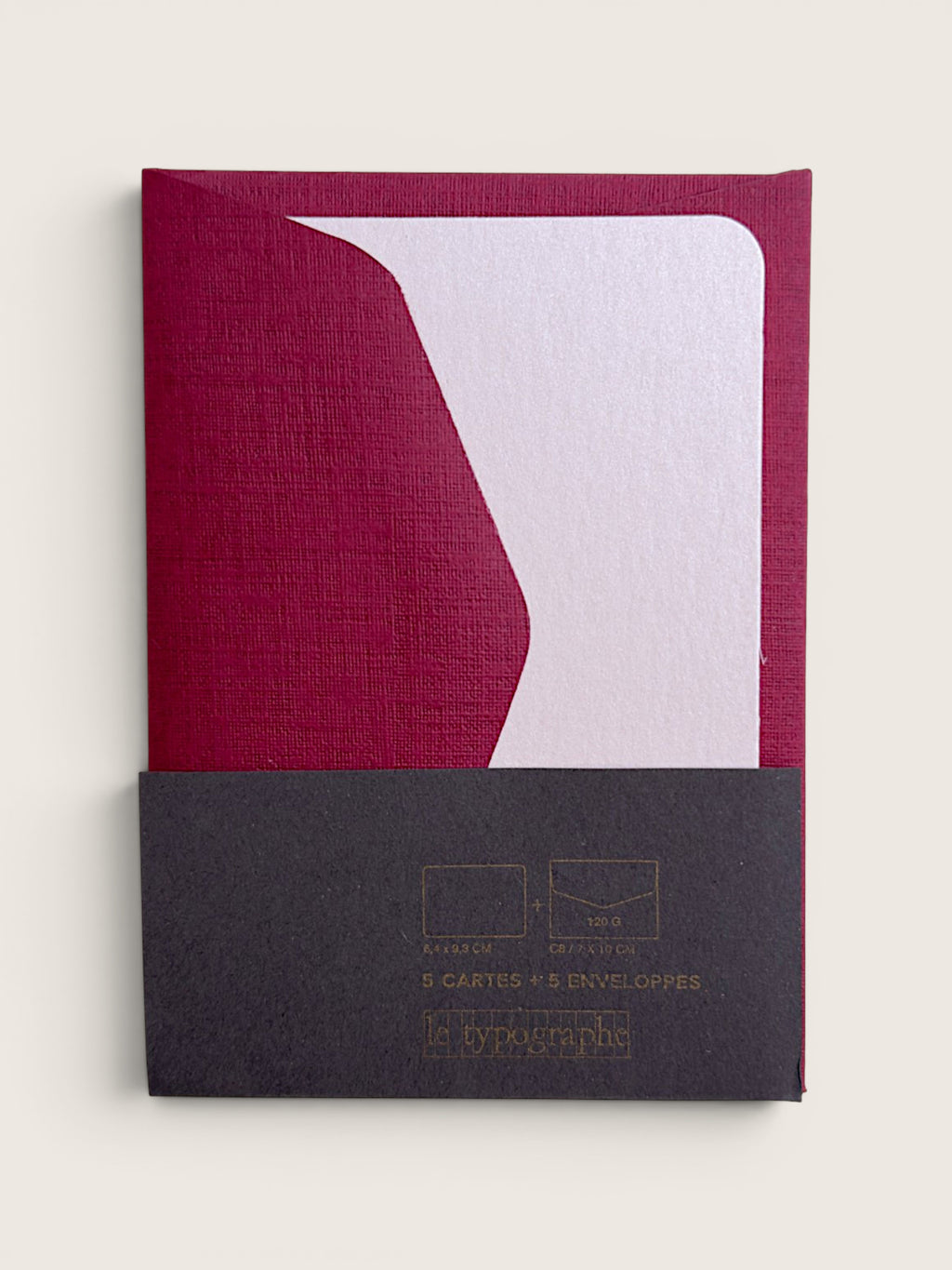 A notebook or journal with a textured deep magenta cover and a white geometric accent, featuring a dark charcoal paper sleeve at the bottom with gold foil text.