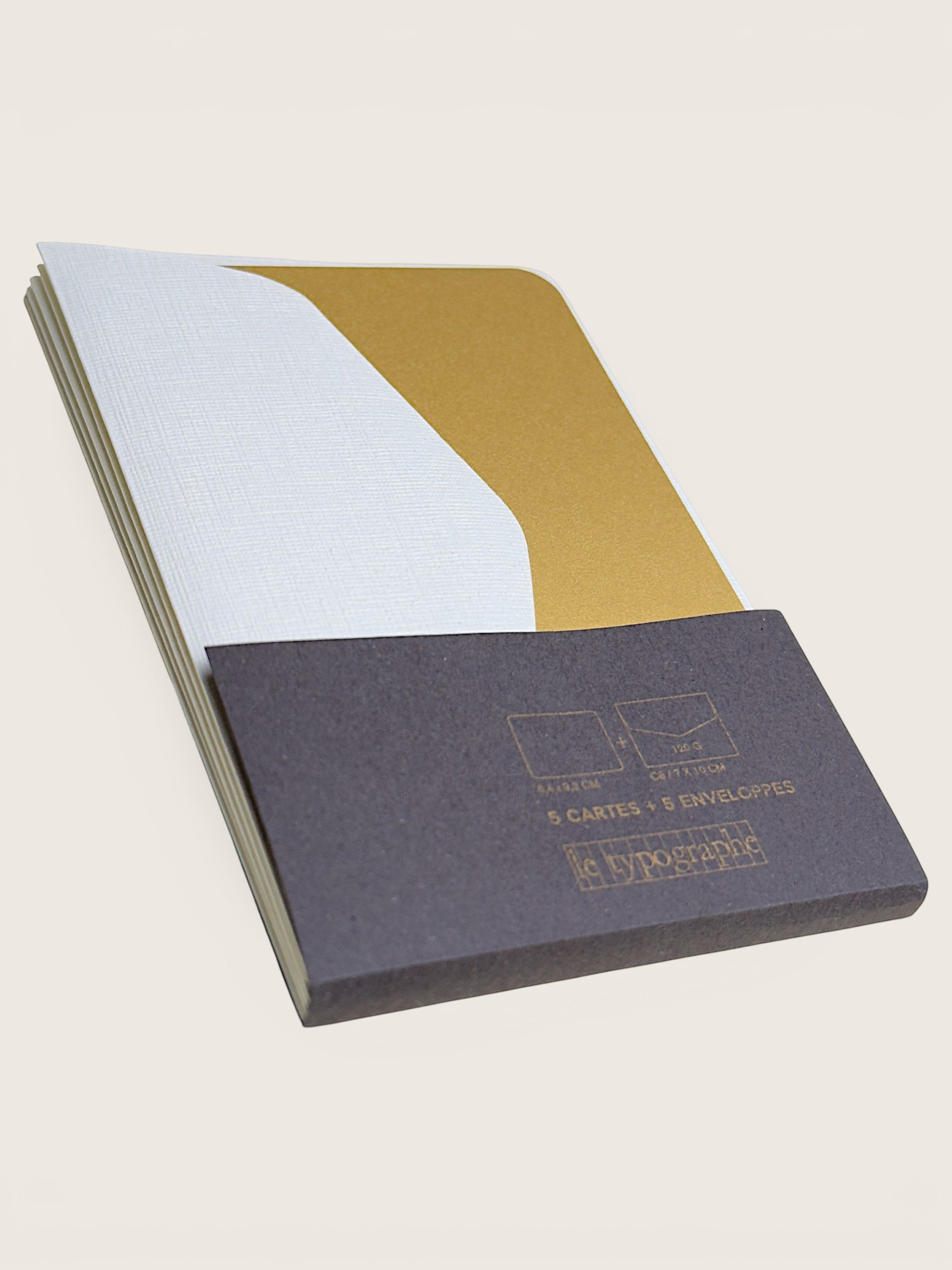 A set of five card and five envelope sets from the brand Enclosure, featuring a dark gray textured cover with gold foil printing, a white envelope set, and a gold card set, displayed on a light beige background.