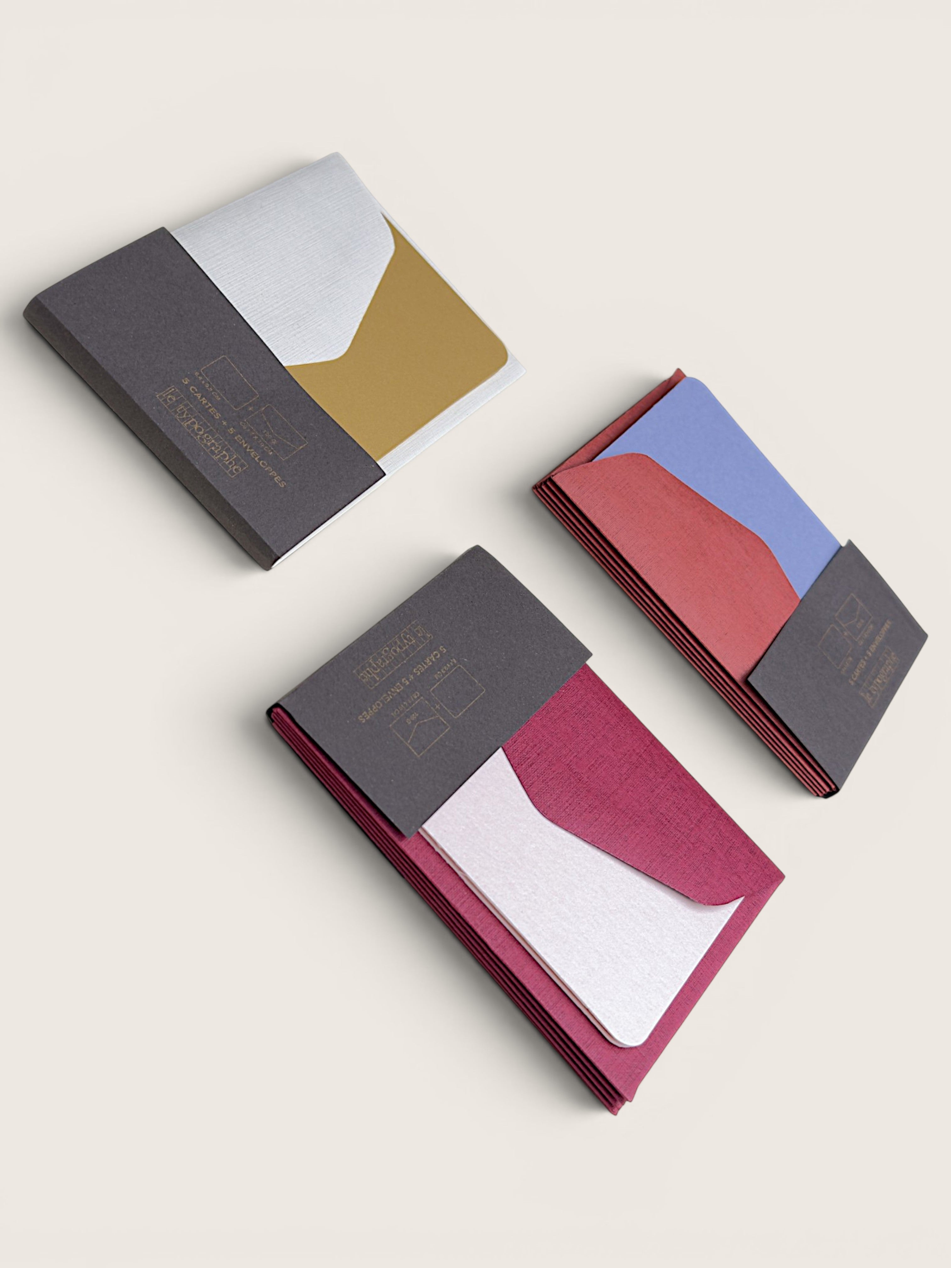 Three slim wallet-style card cases arranged on a light warm beige background. Each case has a folded paper cover with geometric color blocks and embossed branding text.