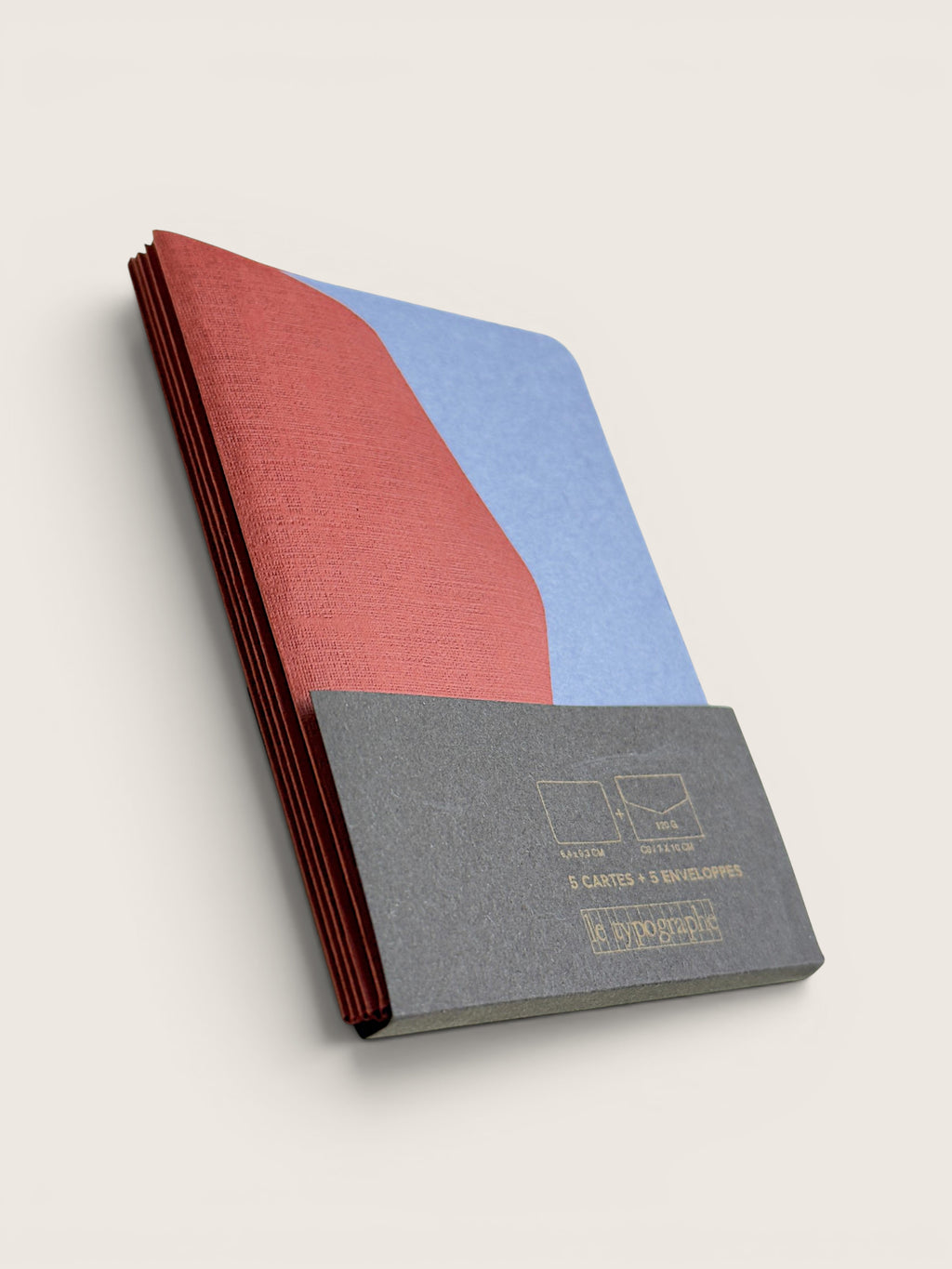 A hardcover notebook or journal shown at an angle on a light beige background. The cover features a color-blocked design with a textured red section on top, a light blue section on the right, and a dark gray textured lower section with gold foil text that reads '3 CARTES x 3 REVISIONS' and 'L'ESPACE DES IDEES'.
