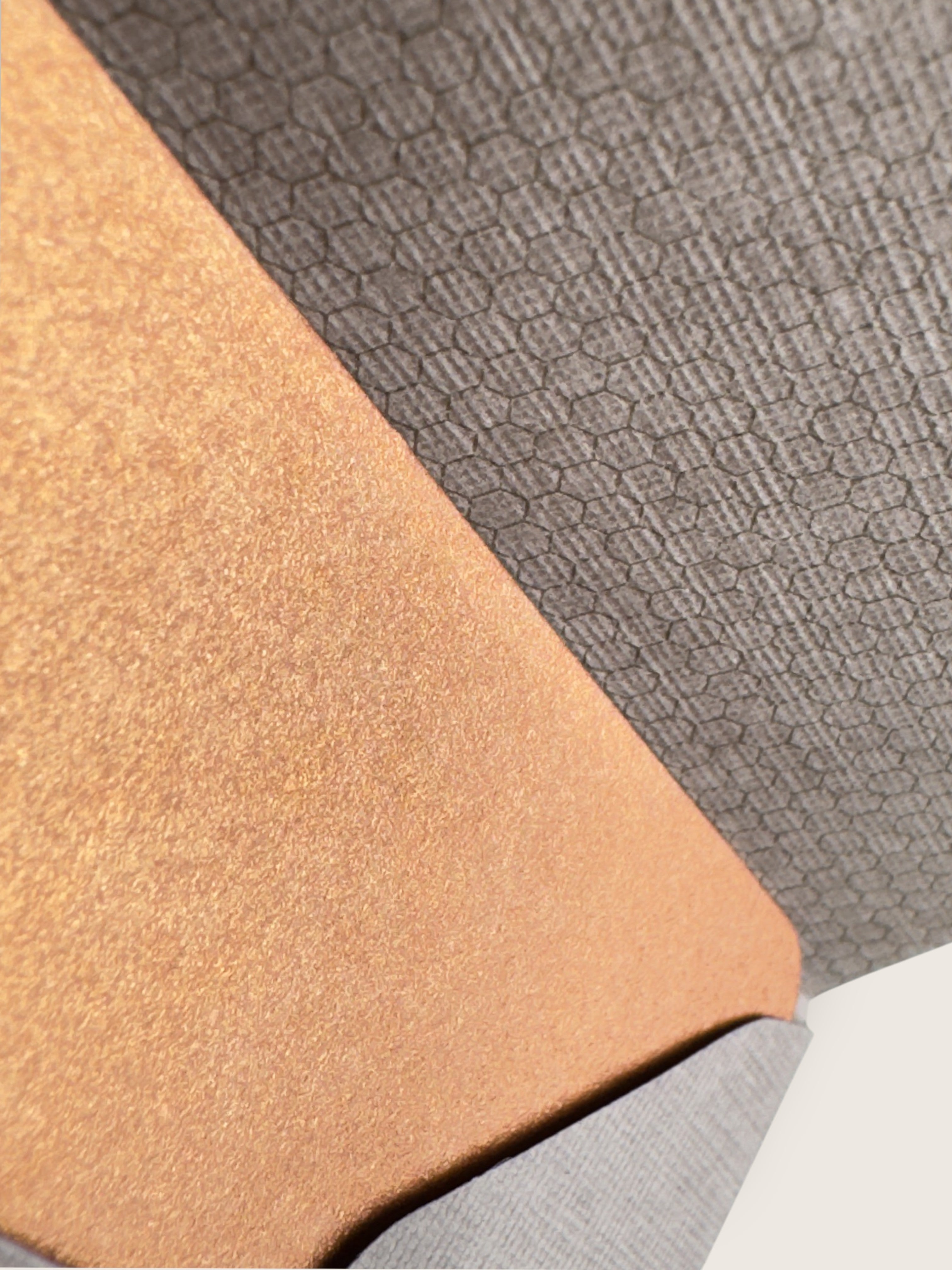 Close-up of two textured material swatches, one in warm tan/camel and one in dark gray with a subtle geometric pattern, overlapping diagonally.