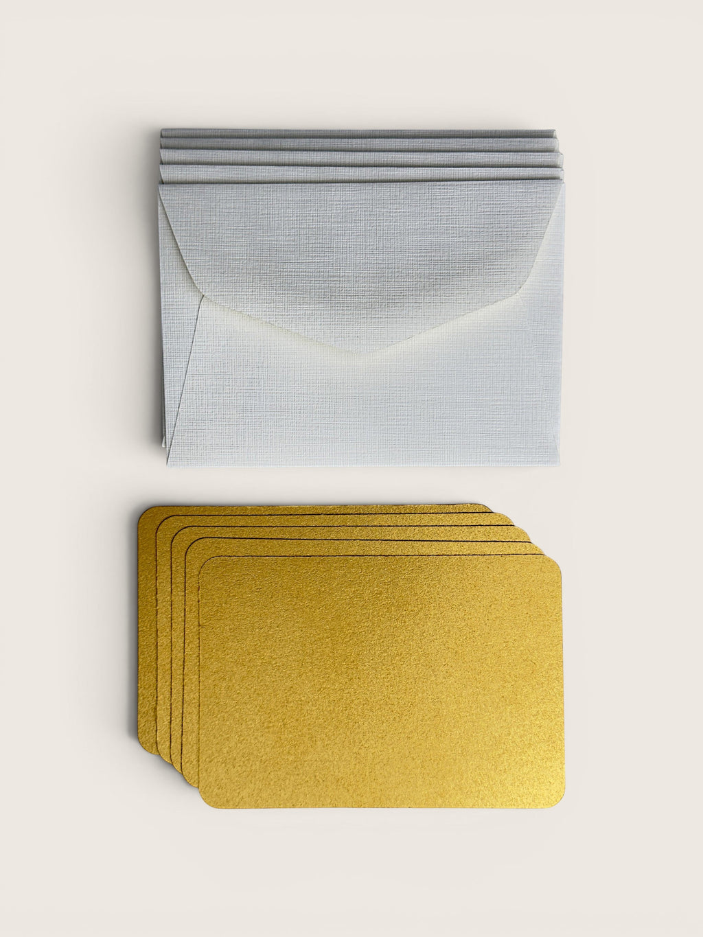 A set of textured white envelopes stacked on top of matching gold metallic cards, arranged neatly on a light beige background.