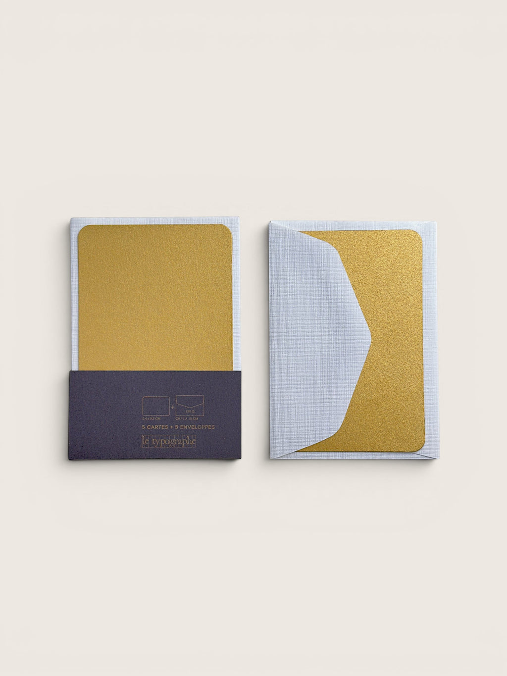 Two small rectangular cards or envelopes with a textured gold center panel and pale blue edges, shown on a plain warm off-white background. One card is closed with a dark navy lower section and printed text, and the other is open to show the pale blue textured side.