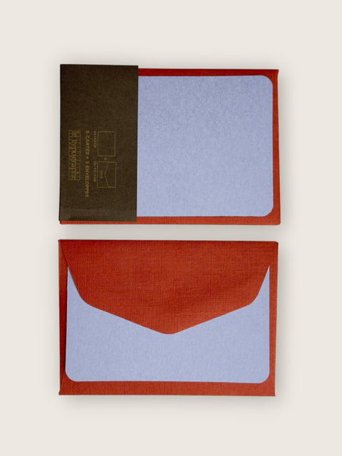 A square pocket notebook with a textured linen cover in terracotta red, featuring a light blue paper insert and a dark brown band with gold text on the left side. The top half shows the notebook closed with the band attached, and the bottom half shows it open with the flap folded down.