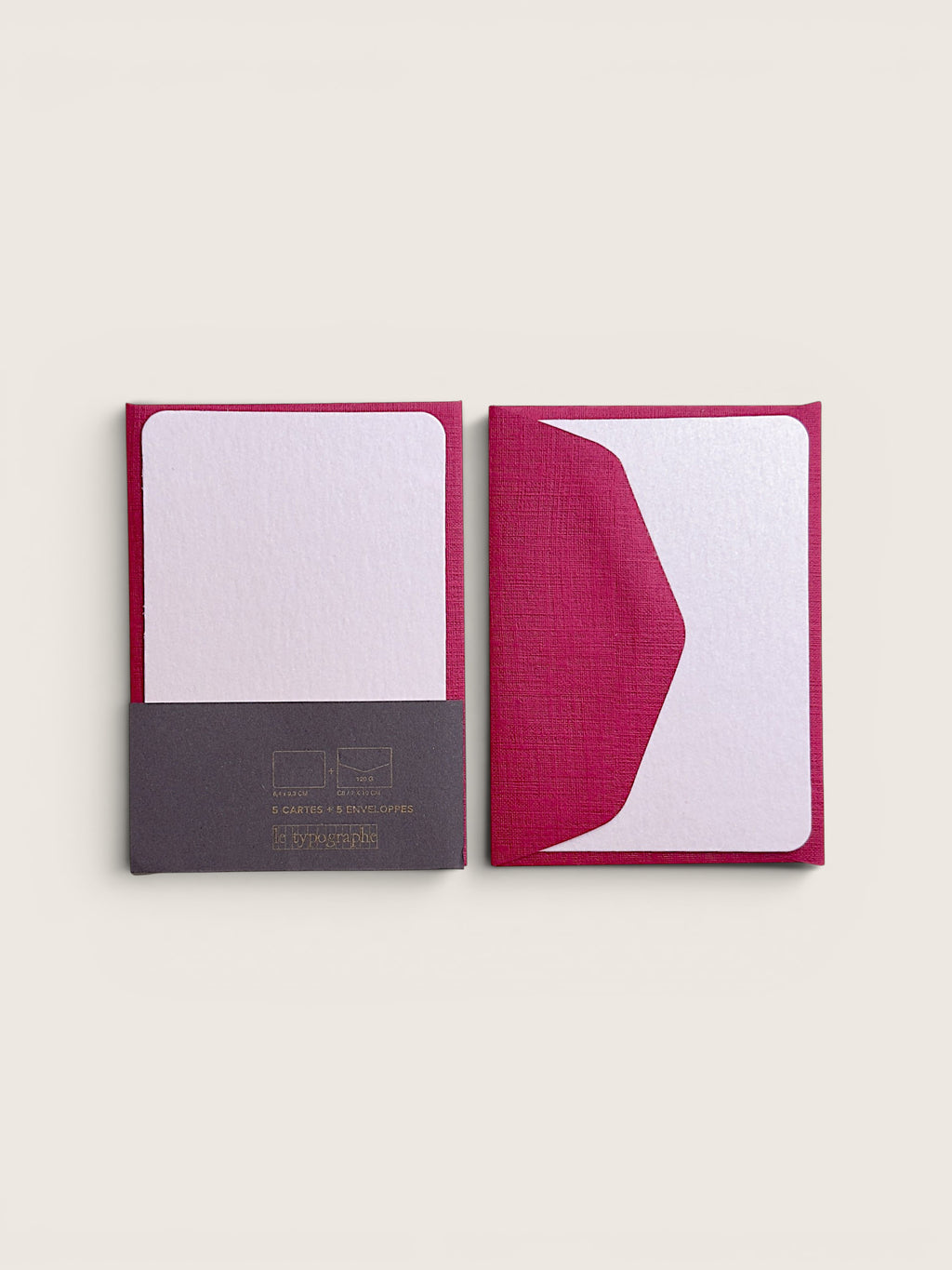 Two slim notebooks or journals with textured covers in a deep magenta pink, shown side by side on a light beige background. The left notebook is open, revealing a white interior page and a dark gray paper sleeve at the bottom with gold text.