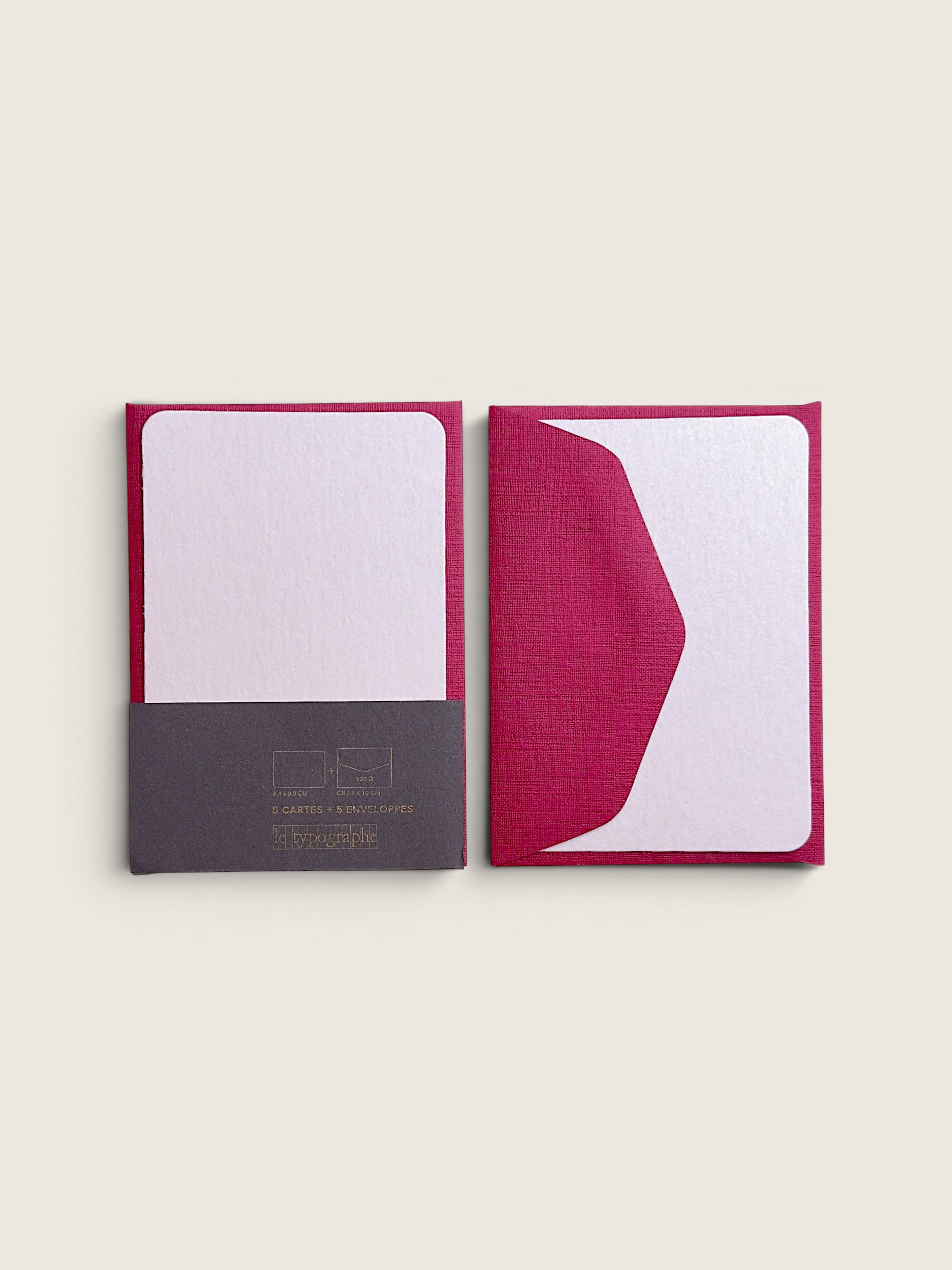 Two slim notebooks or journals with textured covers in a deep magenta pink, shown side by side on a light beige background. The left notebook is open, revealing a white interior page and a dark gray paper sleeve at the bottom with gold text.
