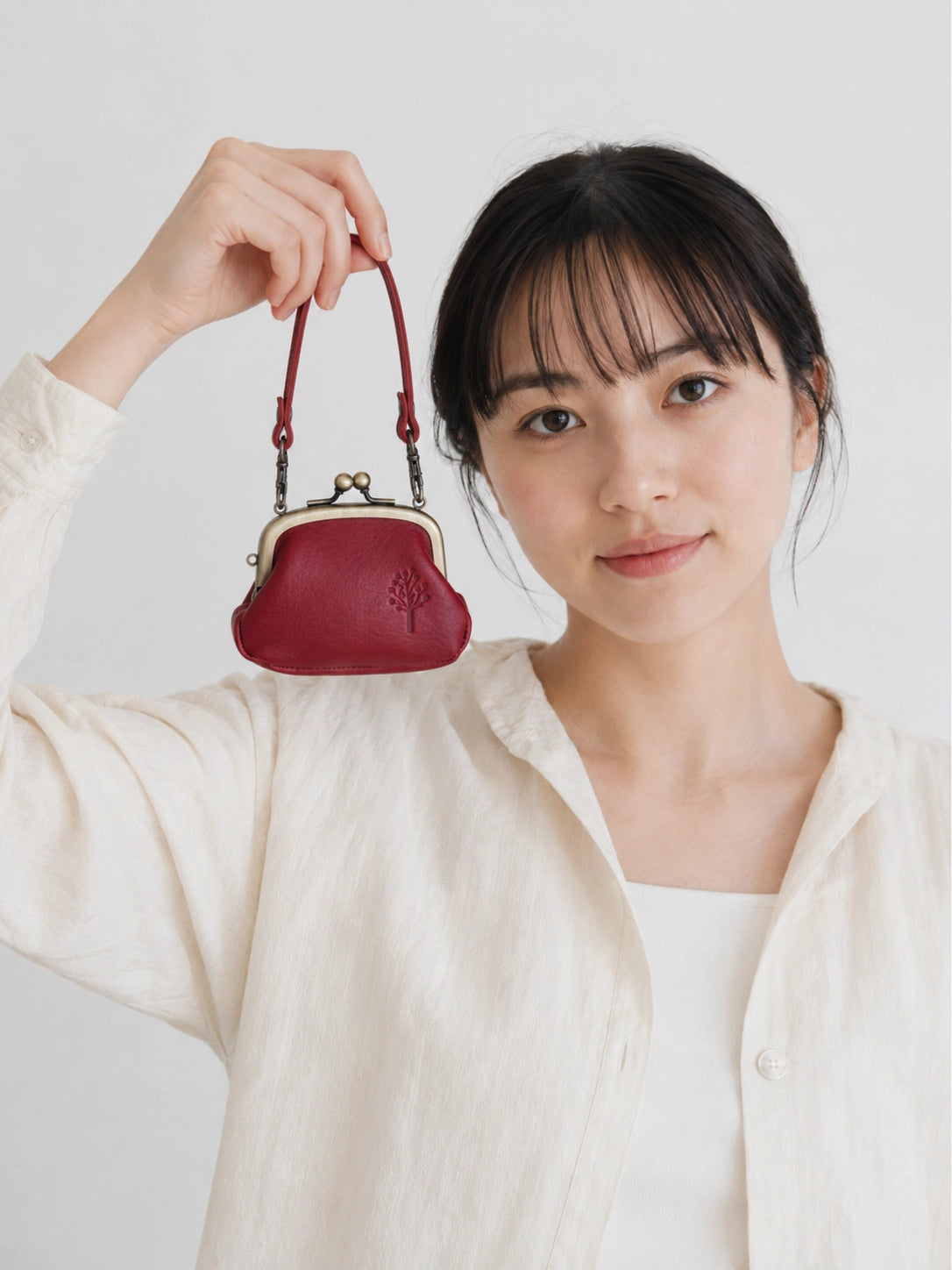 A woman in a cream-colored linen shirt holds up a small, deep red kiss-lock handbag with a matching red strap against a plain light gray background.