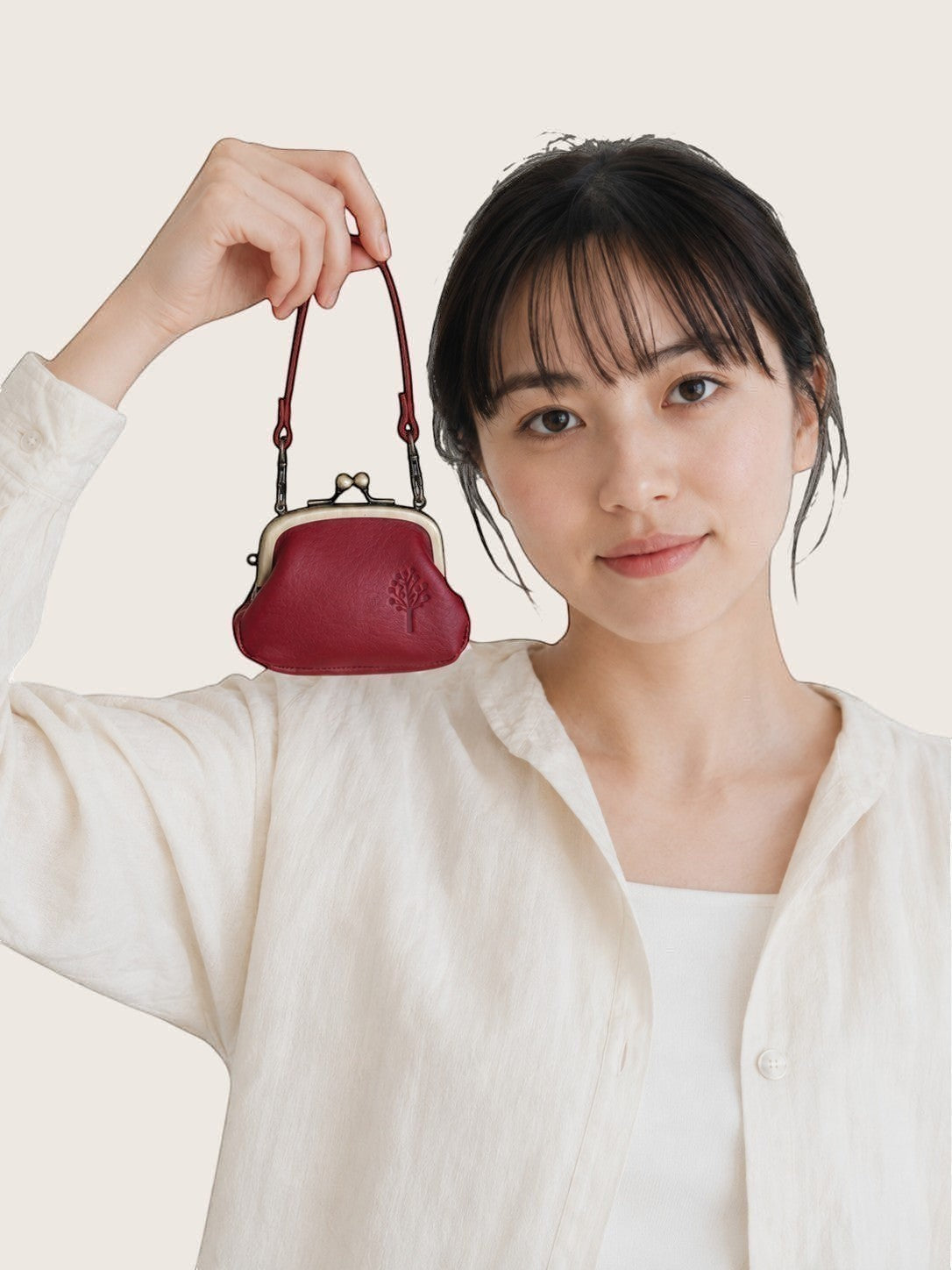 A woman in a white linen shirt holds up a small, dark red leather kiss-lock handbag with a matching shoulder strap against a light beige background.