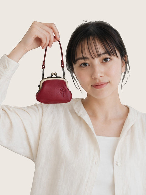 A woman in a white linen shirt holds up a small, dark red leather kiss-lock handbag with a matching shoulder strap against a light beige background.