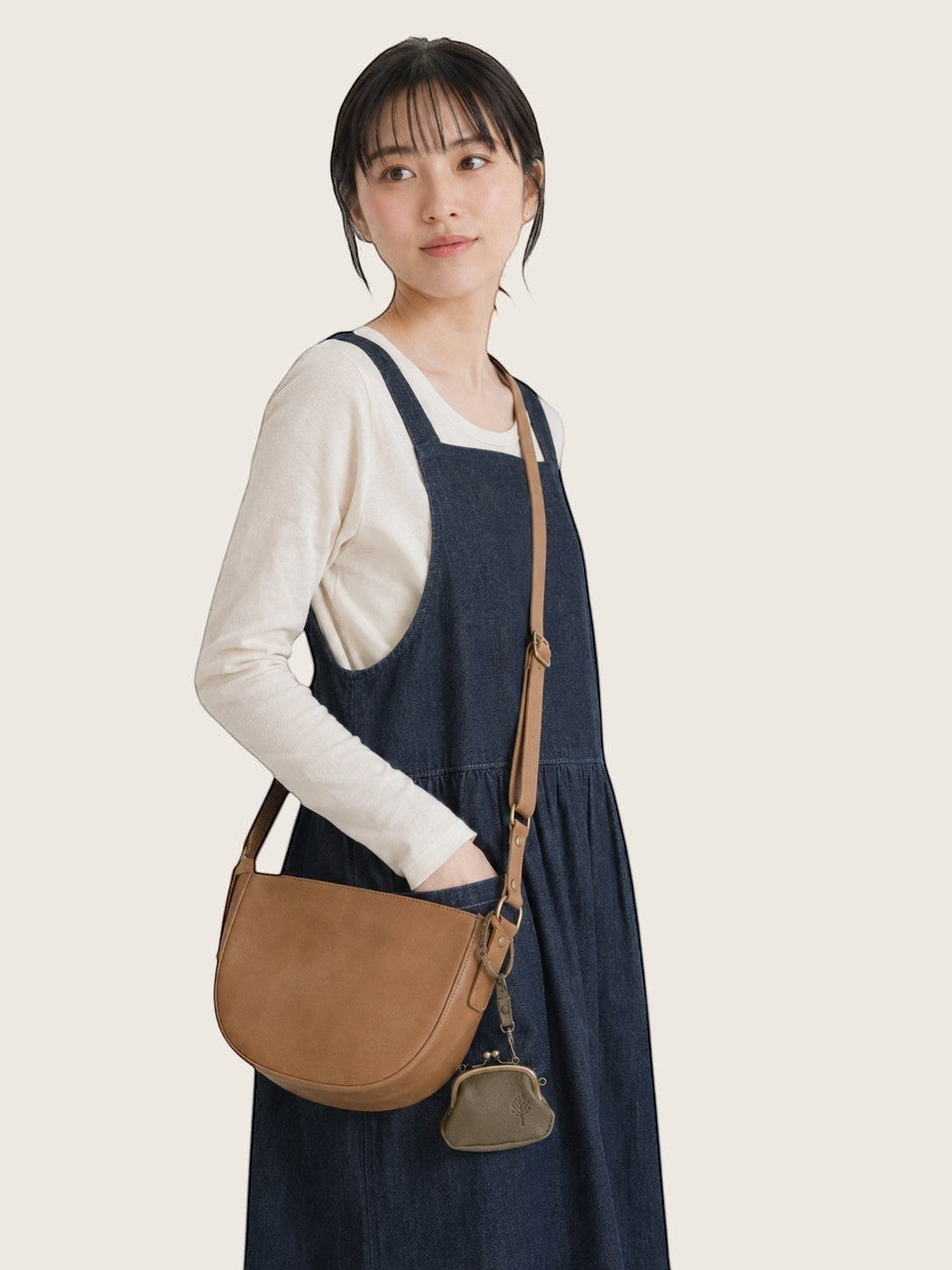 A woman wearing a navy blue denim pinafore dress over a white long-sleeve shirt, carrying a tan leather crescent-shaped shoulder bag with a small matching pouch.