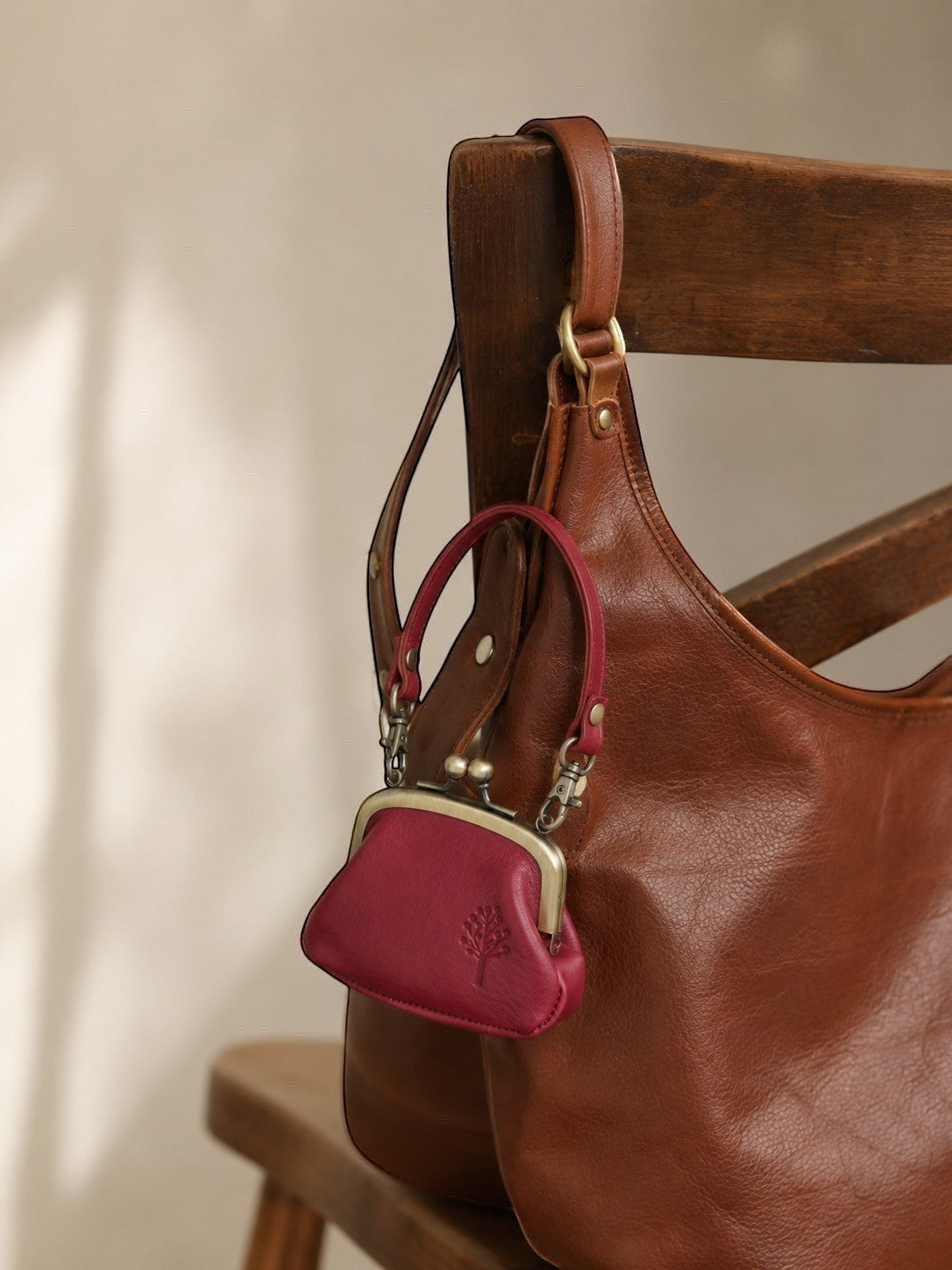 A small magenta leather kiss-lock purse with a silver-tone metal frame hangs from the strap of a large brown leather hobo bag, styled on a wooden chair.