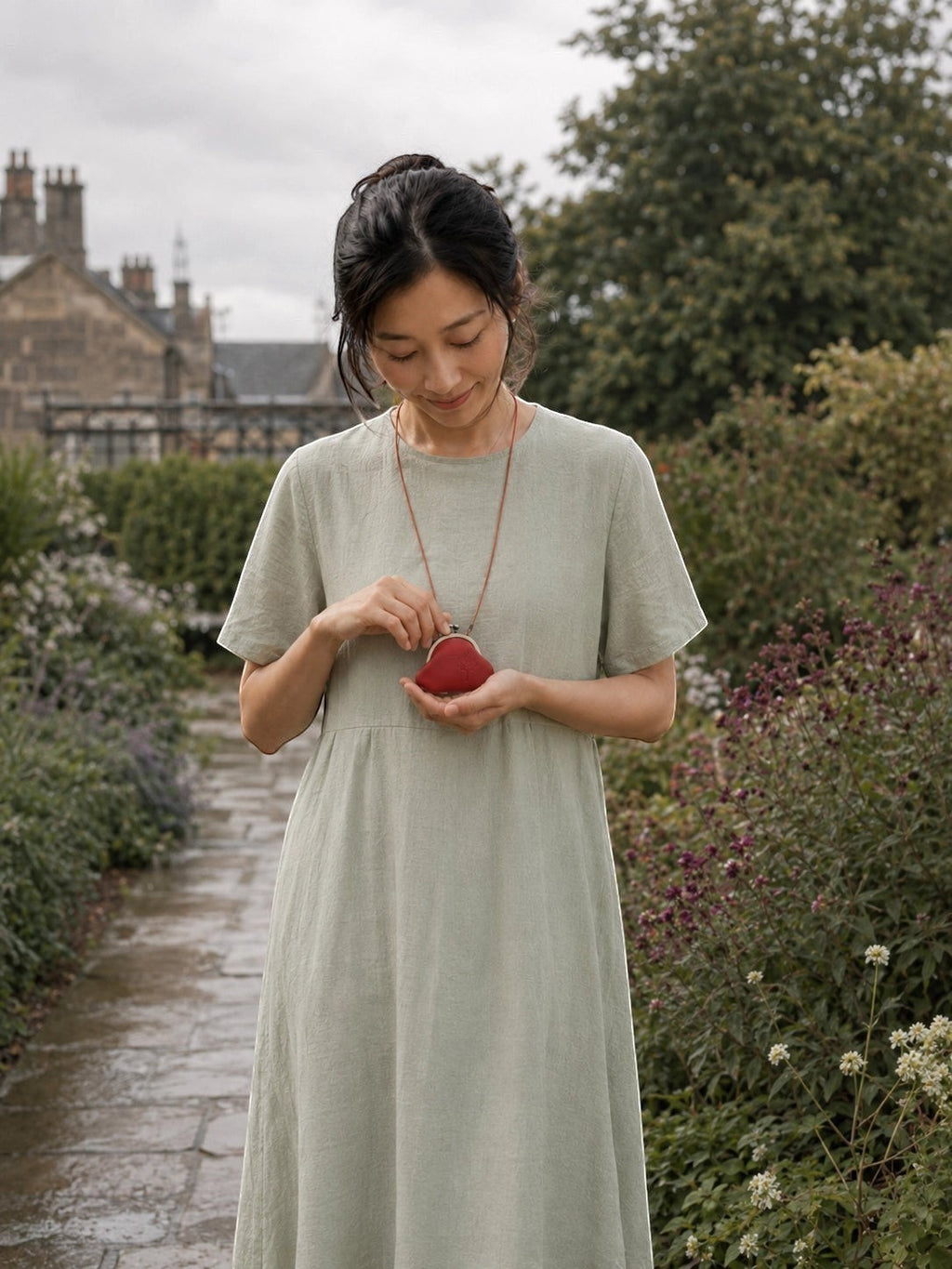 A woman wearing a light sage green linen dress stands in a garden pathway, holding a small red coin purse.