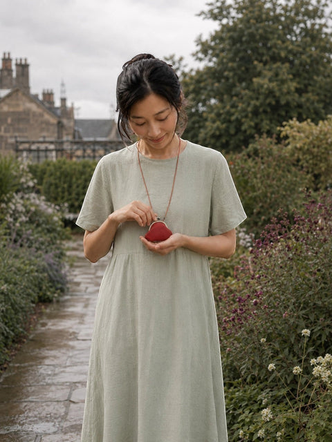 A woman wearing a light sage green linen dress stands in a garden pathway, holding a small red coin purse.