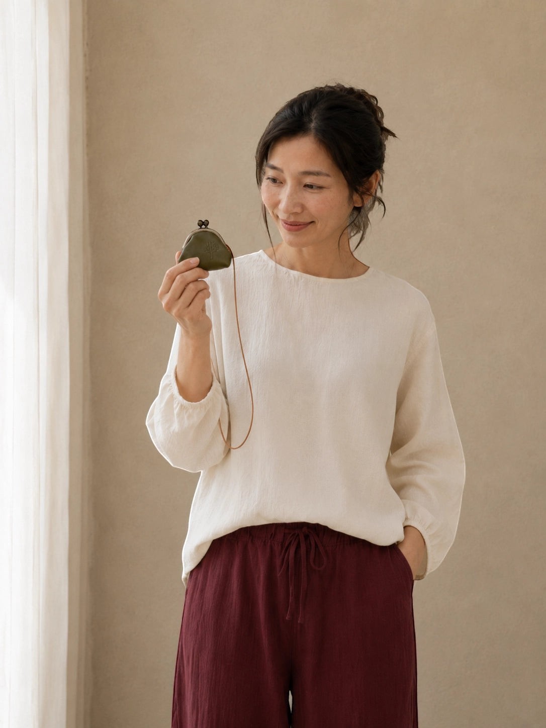 A woman wearing a cream-colored long-sleeve top and dark burgundy drawstring pants, holding a small dark green pouch with a gold clasp against a neutral beige wall.