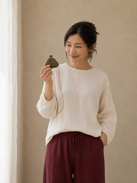 A woman wearing a cream-colored long-sleeve top and dark burgundy drawstring pants, holding a small dark green pouch with a gold clasp against a neutral beige wall.