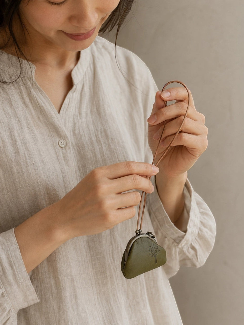 A woman in a light beige linen shirt holds a small, heart-shaped olive green leather pouch with a matching leather strap against a neutral beige background.