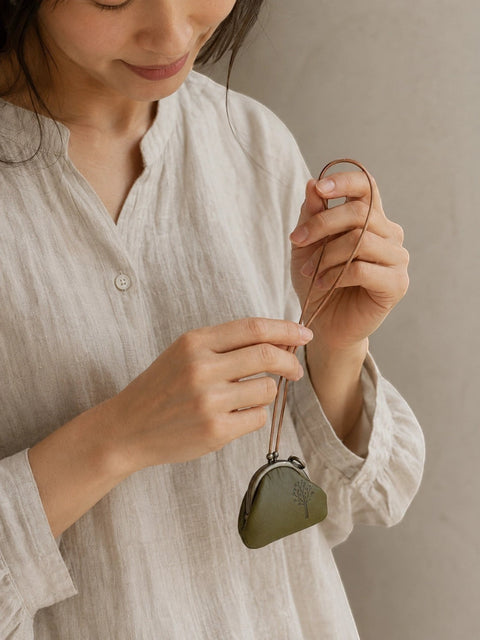 A woman in a light beige linen shirt holds a small, heart-shaped olive green leather pouch with a matching leather strap against a neutral beige background.