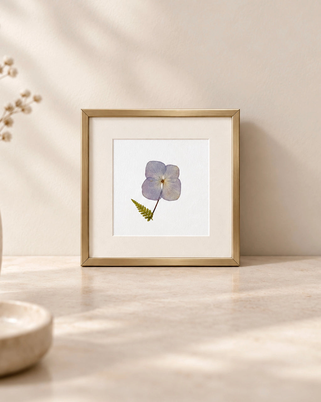 A framed botanical art print featuring a single light pressed Hydrangea botanical flower with a green fern leaf on a white mat, set in a gold frame on a light beige marble surface against a neutral beige wall.